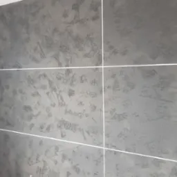 Ściana wykończona szarą, dekoracyjną strukturą z wyraźnymi, nieregularnymi wzorami i białymi fugami, imitująca duże płytki.