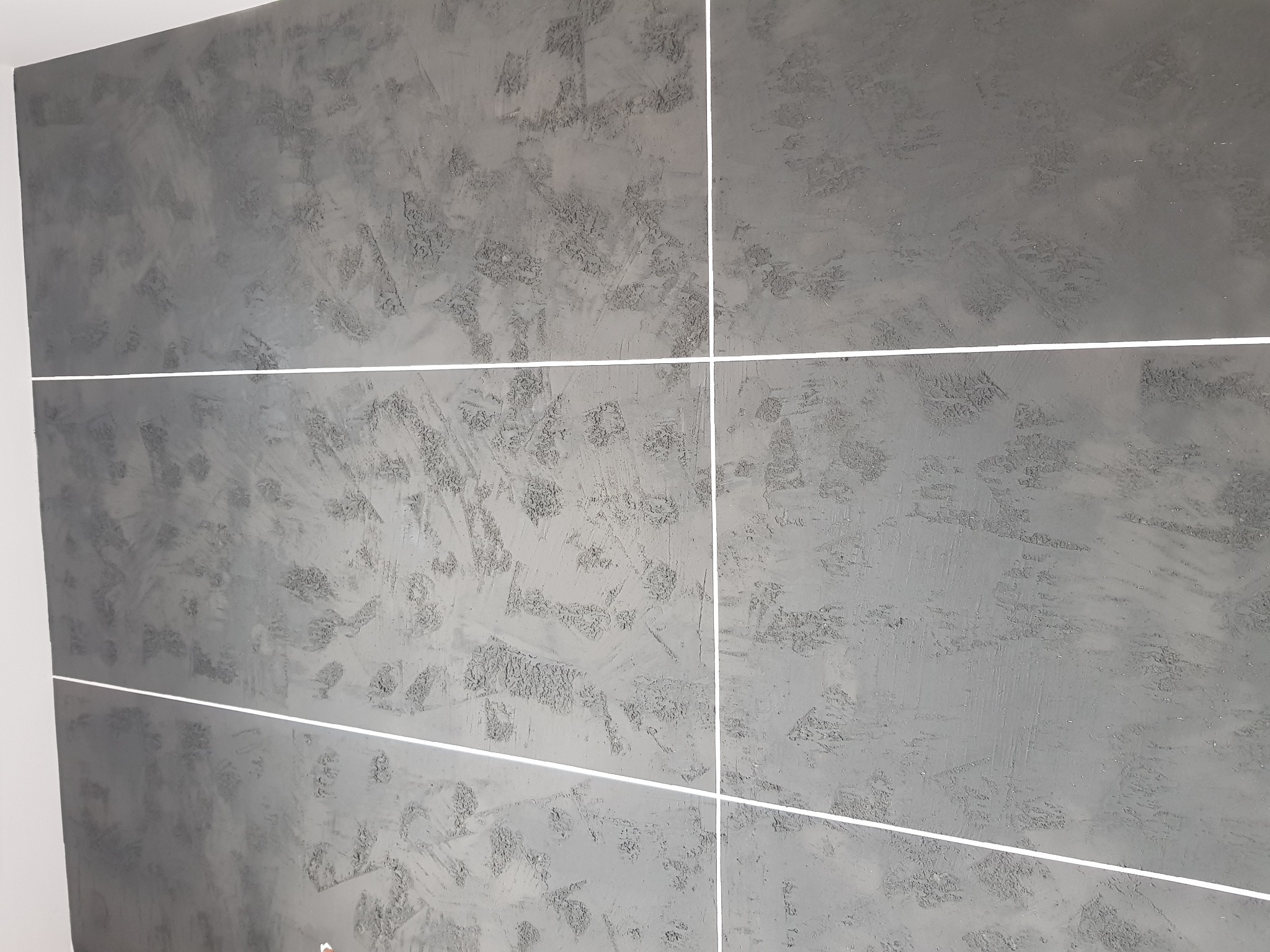 Ściana wykończona szarą, dekoracyjną strukturą z wyraźnymi, nieregularnymi wzorami i białymi fugami, imitująca duże płytki.