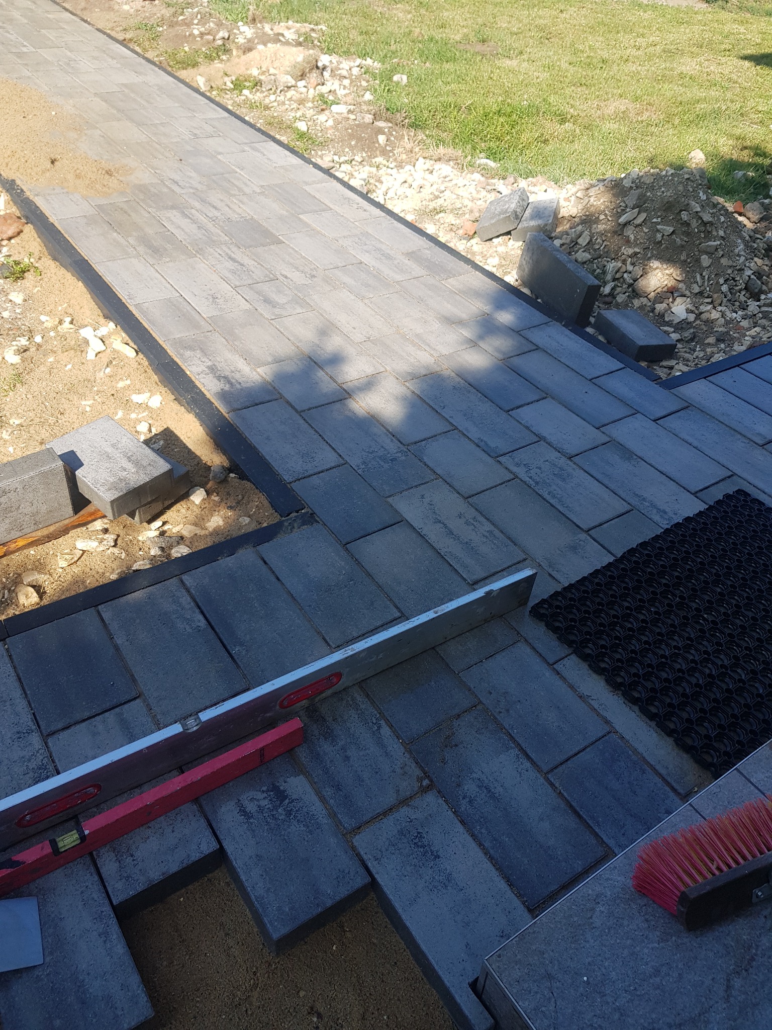 Układanie szarej kostki brukowej na ścieżce ogrodowej, widoczna poziomica, szczotka i mata gumowa, etap prac ziemnych z podsypką piaskową.