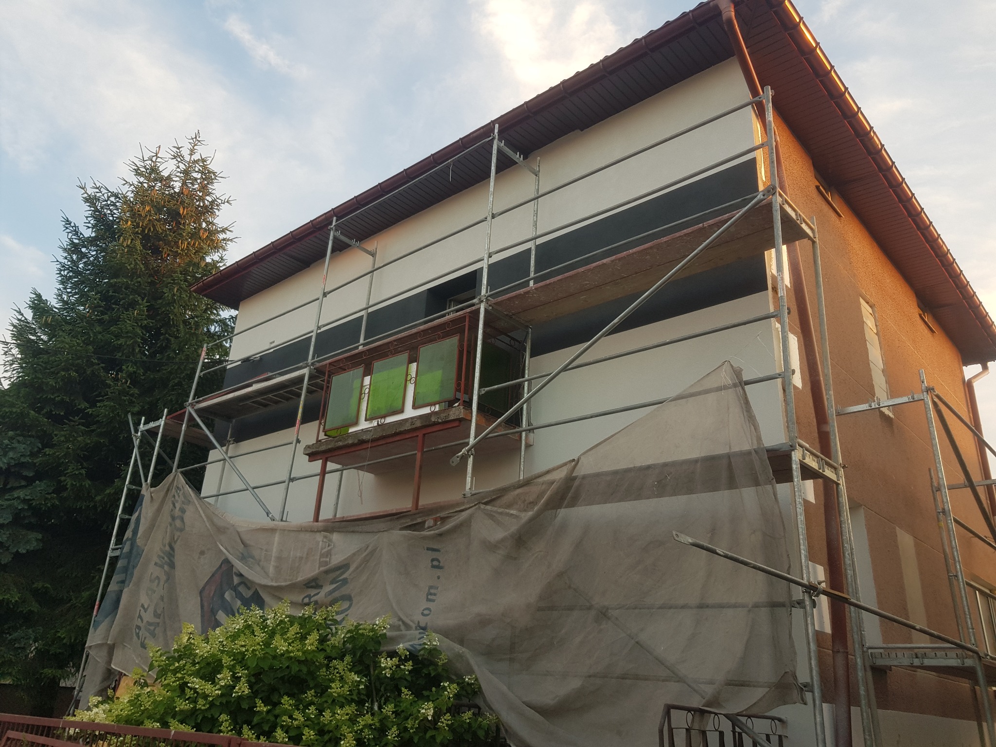 Dom jednorodzinny w trakcie prac elewacyjnych, widoczne rusztowania i plandeka ochronna z logo producenta materiałów budowlanych.