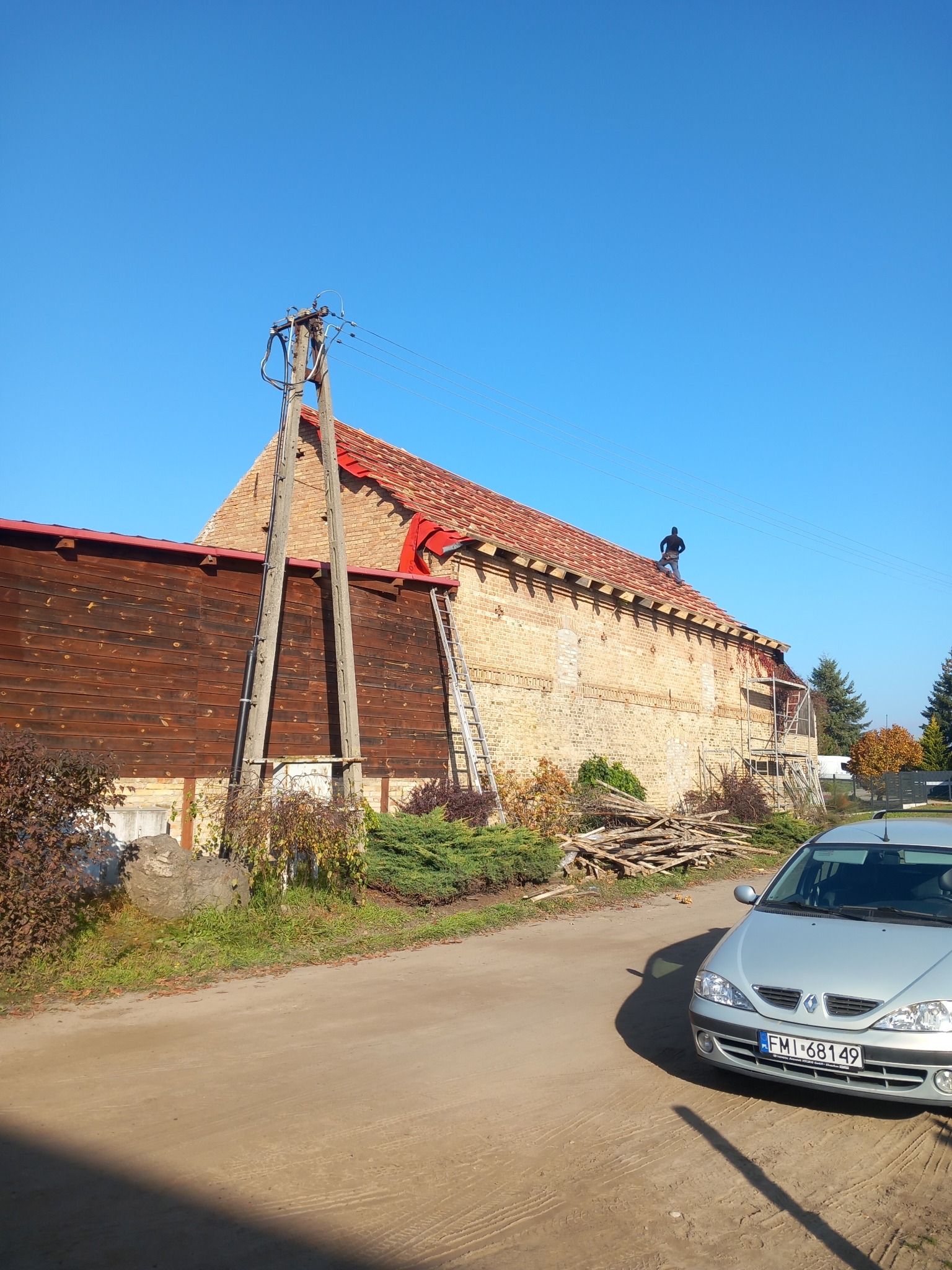 Murarz na dachu ceglanego budynku z drewnianą dobudówką, drabina oparta o ścianę, samochód osobowy w tle.