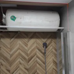 Bojler elektryczny zamontowany na półce w łazience, widoczne rury, grzejnik drabinkowy, ściana z płytek ułożonych w jodełkę oraz kabina prysznicowa