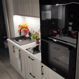 Zbigniew Zagórny - Nowoczesna kuchnia z frontami w odcieniach jasnego drewna, czarnym zlewem i baterią, płytą indukcyjną oraz piekarnikiem z wyświetlaczem, dekorowana kwiatami i pomarańczowymi ozdobami.