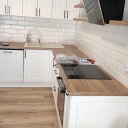 Zbigniew Zagórny - Nowoczesna, jasna kuchnia z białymi frontami szafek, drewnianym blatem, płytą indukcyjną i białymi kafelkami na ścianie, oświetlona lampą sufitową.