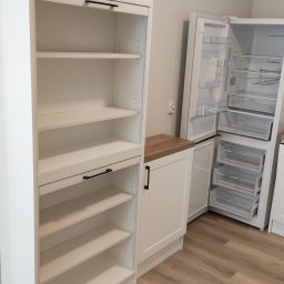 Zbigniew Zagórny - Biała zabudowa kuchenna z szafką z półkami i szafką z blatem z drewnopodobną okleiną, obok otwarta lodówka z widocznymi półkami i szufladami.