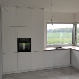 Zbigniew Zagórny - Jasna, minimalistyczna kuchnia z białymi szafkami, czarnym piekarnikiem i drewnianym blatem, widok na zielone pole za oknem, błyszcząca podłoga z płytek.