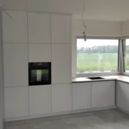 Jasna, minimalistyczna kuchnia z białymi szafkami, czarnym piekarnikiem i drewnianym blatem, widok na zielone pole za oknem, błyszcząca podłoga z płytek.