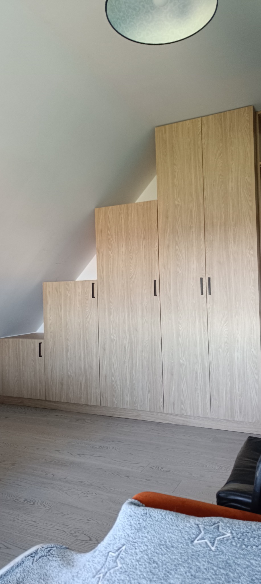 Zabudowa wnęki pod skosem z jasnymi szafami z drewnopodobnym dekorem i czarnymi uchwytami, widok z dołu z fragmentem sofy z przodu.