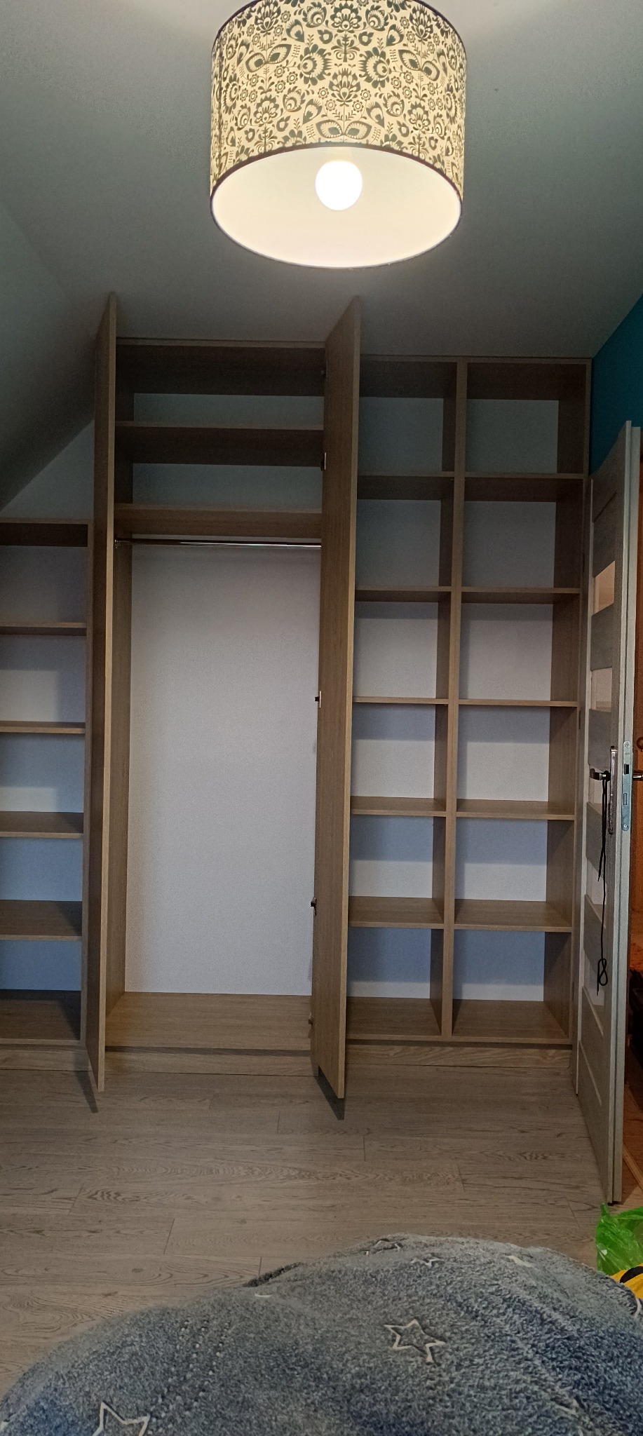 Otwarta, jasna szafa wnękowa z półkami i drążkiem na ubrania, oświetlona lampą z wzorem, na poddaszu.