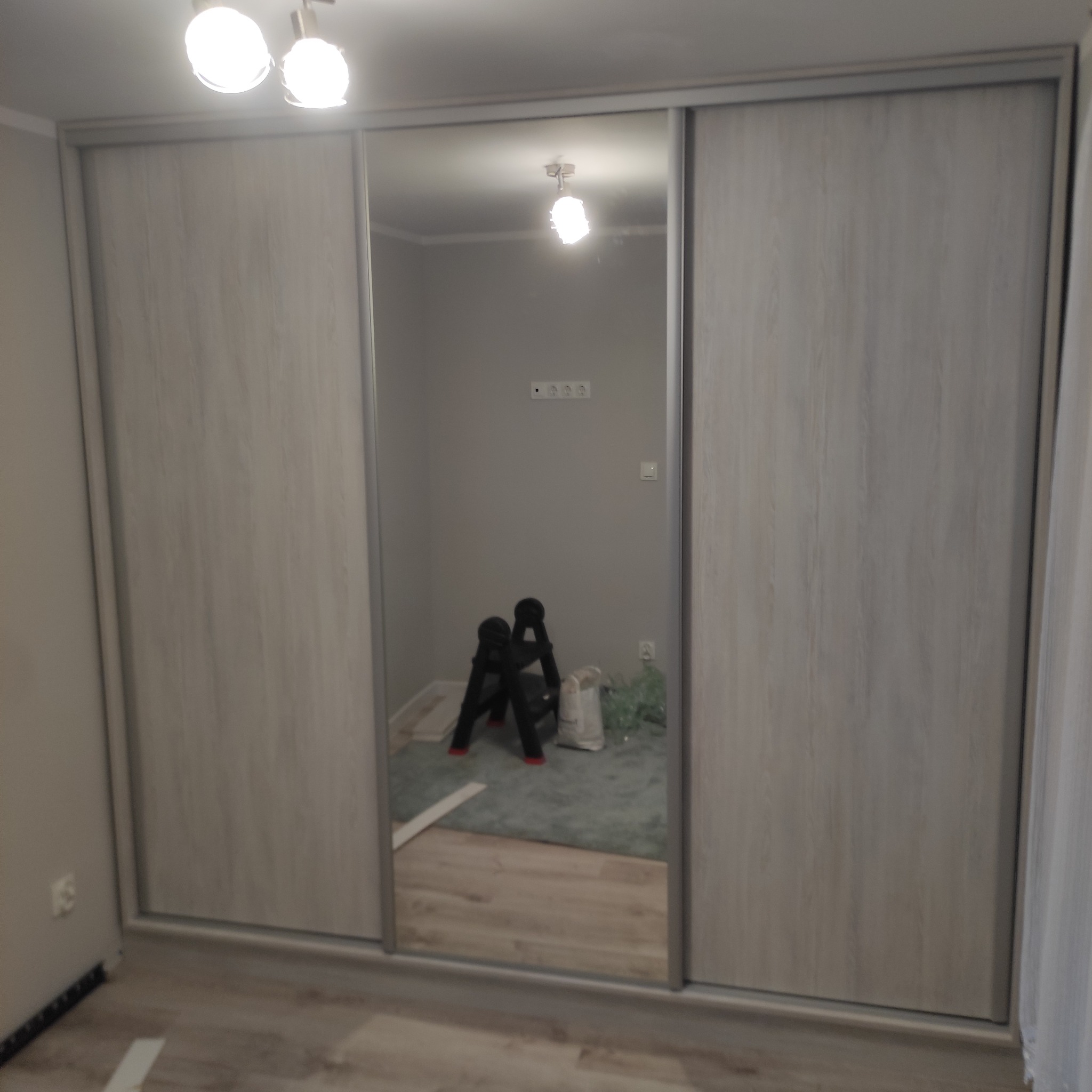 Trzydrzwiowa szafa przesuwna z lustrem w centralnej części, widoczna drabina, materiały budowlane i gniazdka elektryczne w tle.