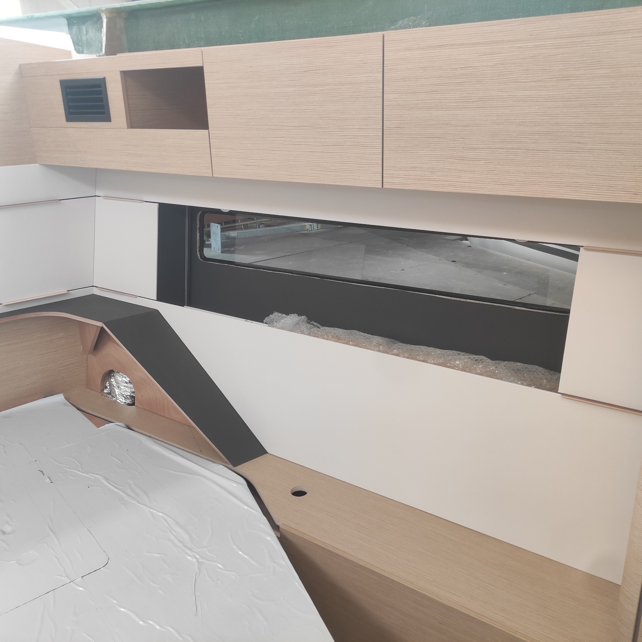 Wnętrze jachtu w trakcie wykończenia: jasne drewniane szafki z wentylacją, okno z widokiem na zewnątrz, elementy konstrukcyjne w trakcie montażu, zabezpieczone folią.