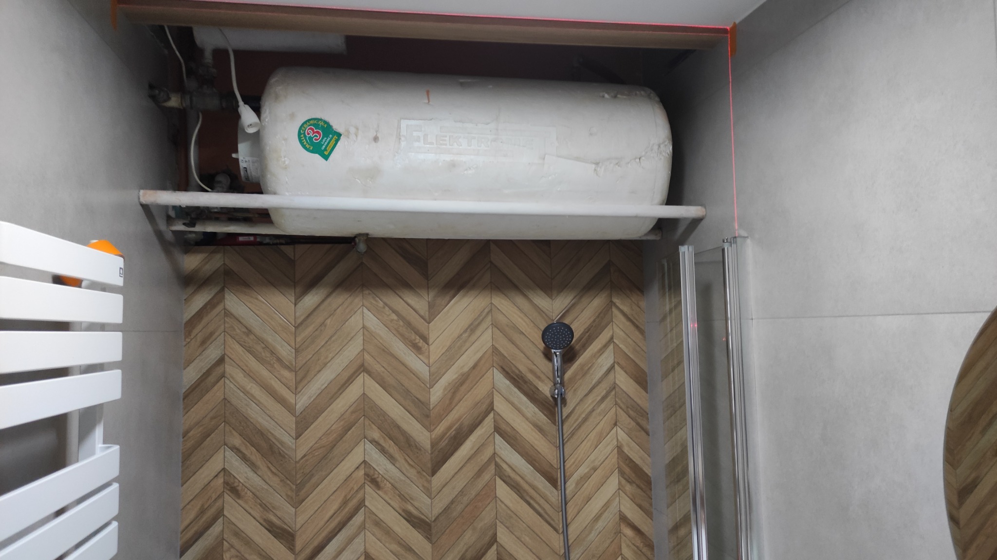 Bojler elektryczny zamontowany na półce w łazience, widoczne rury, grzejnik drabinkowy, ściana z płytek ułożonych w jodełkę oraz kabina prysznicowa
