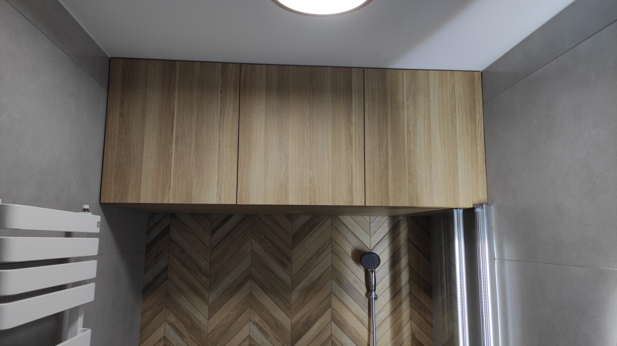 Drewniane szafki wiszące nad prysznicem z mozaikową ścianą w jodełkę, widoczny grzejnik drabinkowy i fragment kabiny prysznicowej.