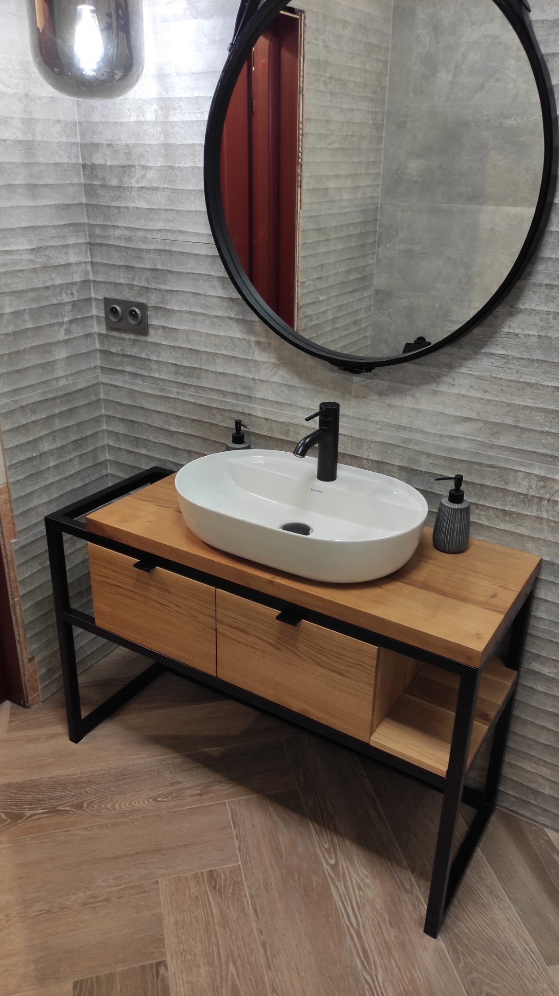 Nowoczesna łazienka z umywalką nablatową na drewnianej szafce z czarną metalową ramą, okrągłe lustro w czarnej ramie i szara ściana z poziomymi paskami.