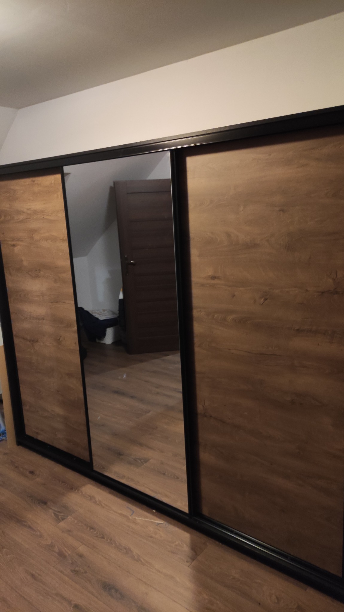 Szafa przesuwna z lustrzanymi drzwiami i okleiną drewnopodobną w pokoju na poddaszu, widoczne odbicie wnętrza.