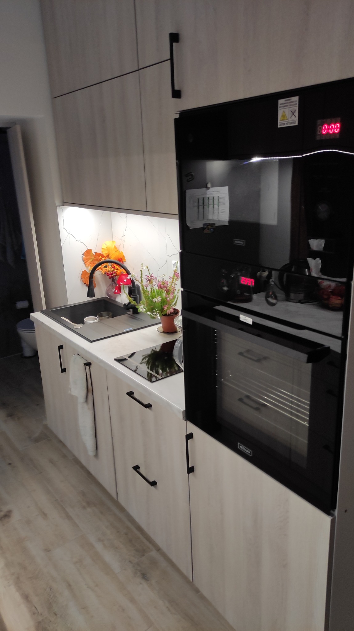 Nowoczesna kuchnia z frontami w odcieniach jasnego drewna, czarnym zlewem i baterią, płytą indukcyjną oraz piekarnikiem z wyświetlaczem, dekorowana kwiatami i pomarańczowymi ozdobami.