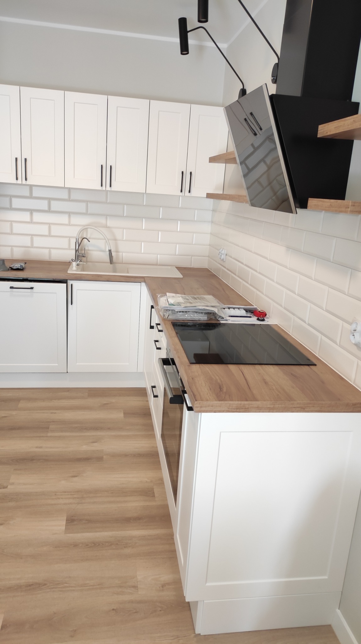 Nowoczesna, jasna kuchnia z białymi frontami szafek, drewnianym blatem, płytą indukcyjną i białymi kafelkami na ścianie, oświetlona lampą sufitową.