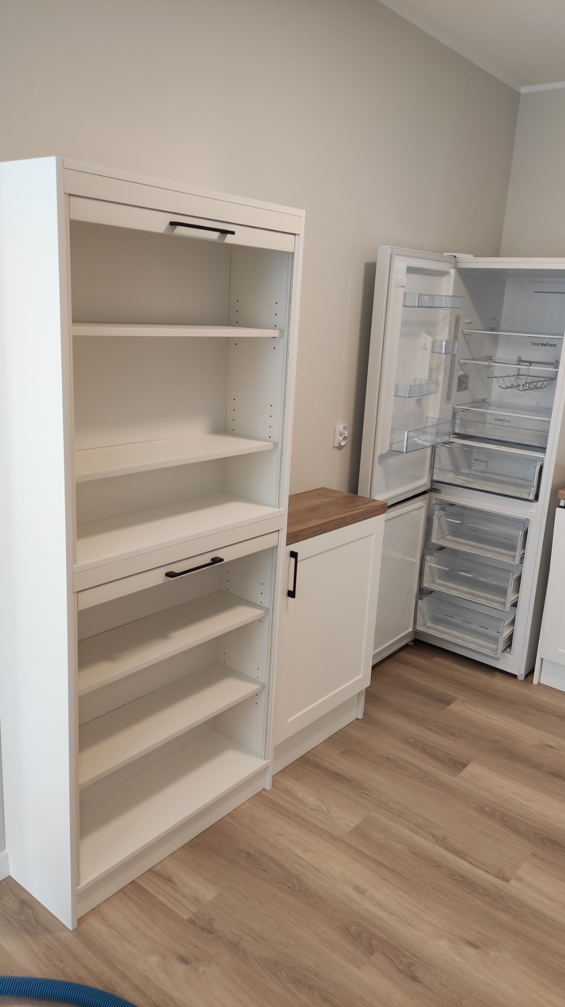 Biała zabudowa kuchenna z szafką z półkami i szafką z blatem z drewnopodobną okleiną, obok otwarta lodówka z widocznymi półkami i szufladami.