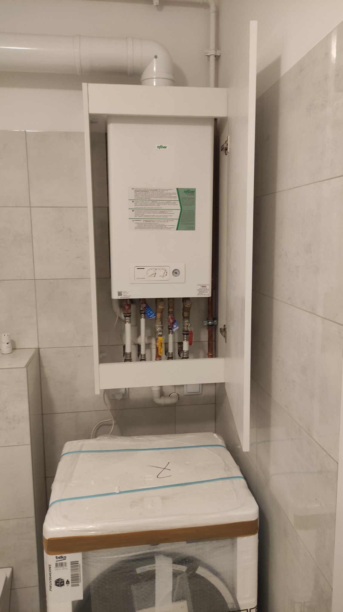 Biały piec gazowy marki Syber w szafce w łazience z otwartymi drzwiczkami, widoczne rury i zawory, poniżej nowa pralka w folii.