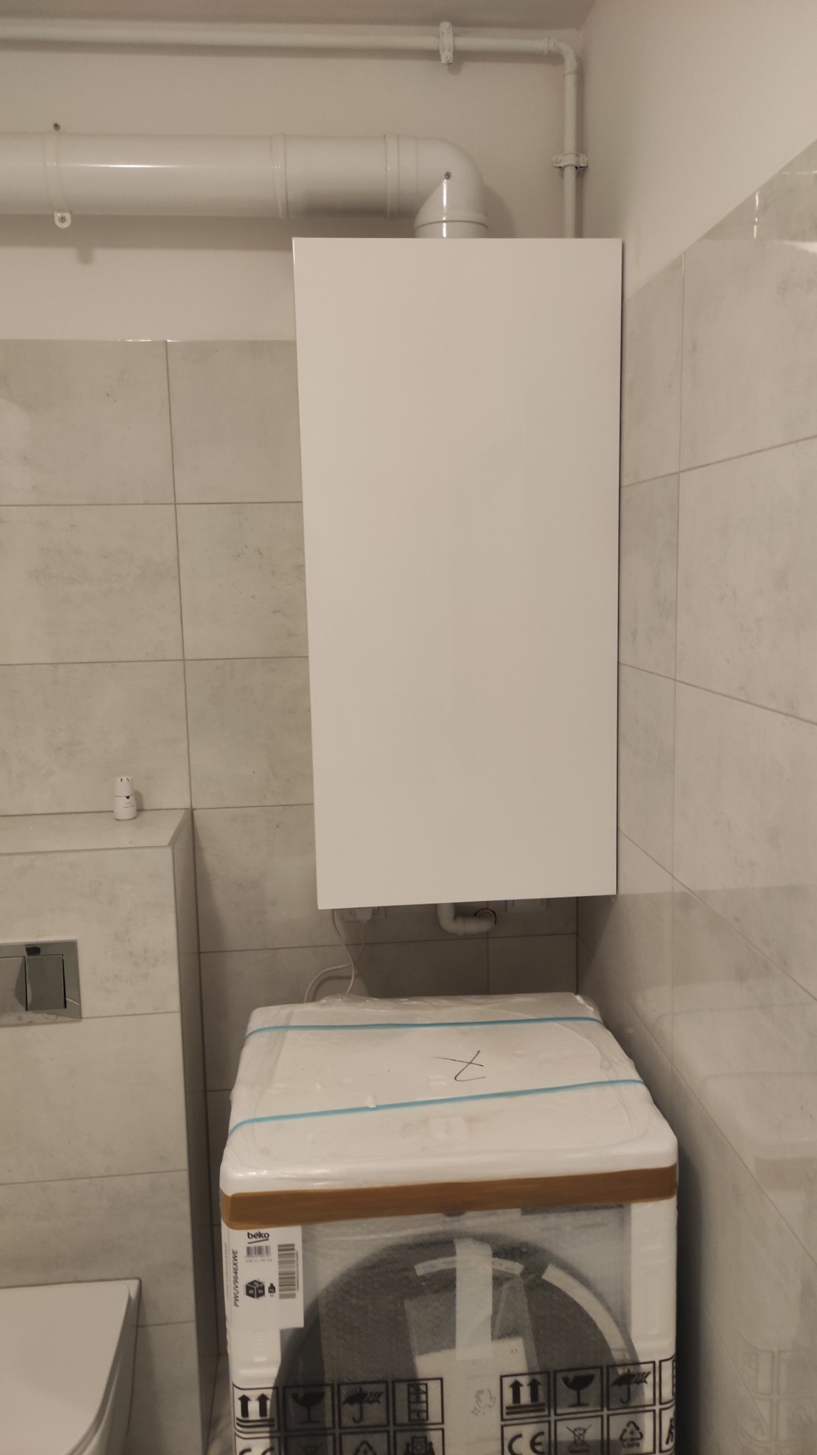 Biały piec gazowy wiszący na ścianie w łazience z beżowymi płytkami, poniżej nowa pralka w fabrycznym opakowaniu.