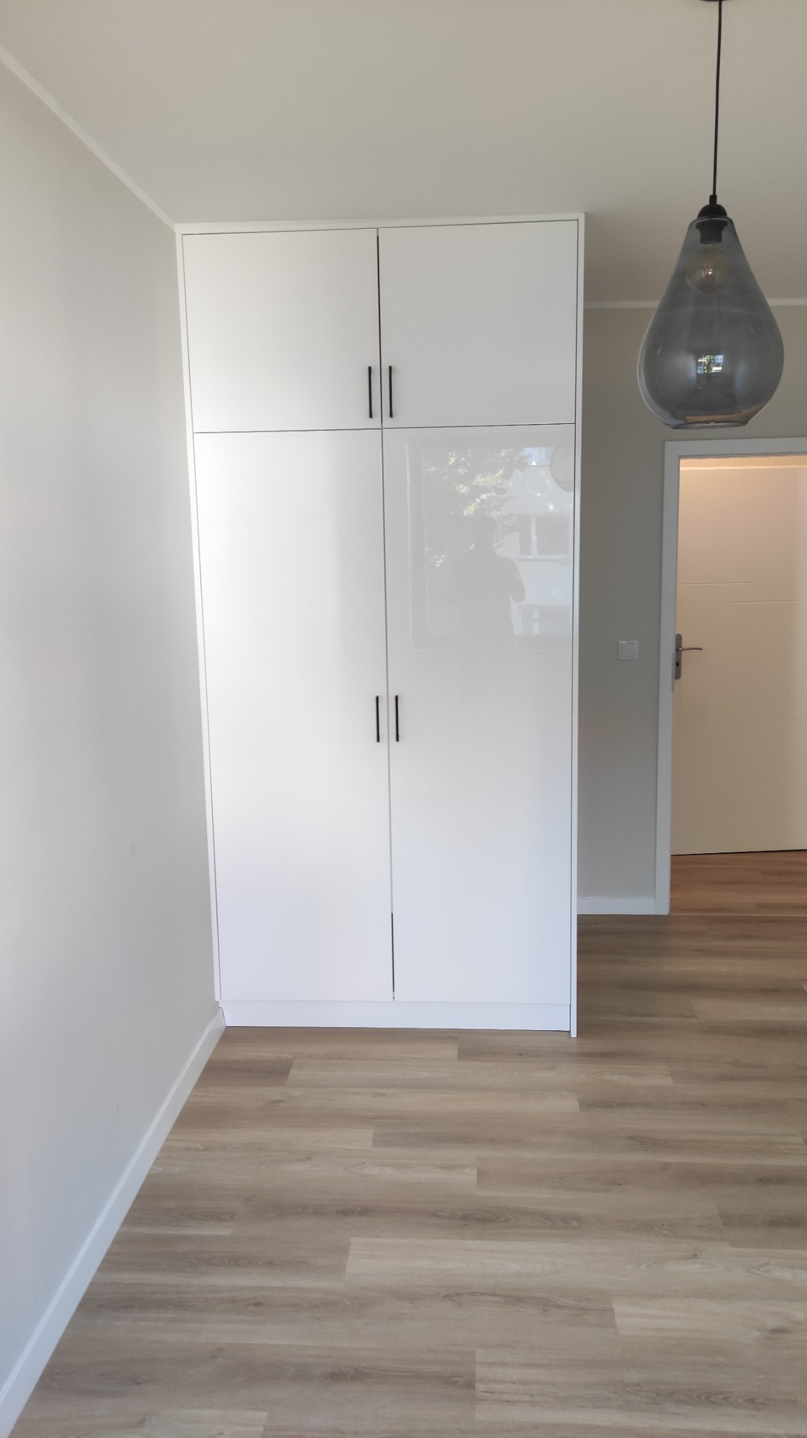 Biała, wysoka szafa z połyskiem i czarnymi uchwytami, dopasowana do sufitu, na tle jasnego wnętrza z panelami podłogowymi i szarą lampą wiszącą.