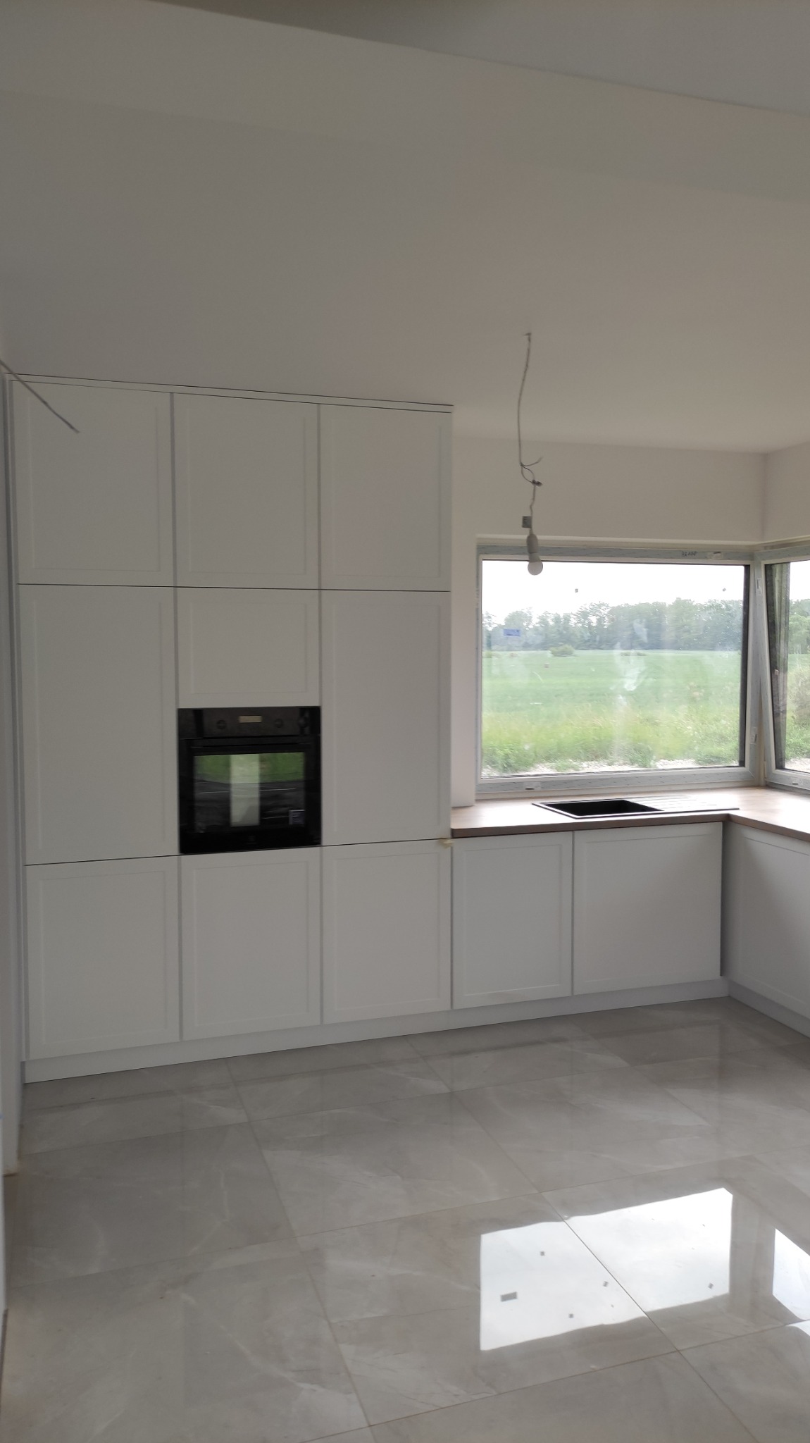 Jasna, minimalistyczna kuchnia z białymi szafkami, czarnym piekarnikiem i drewnianym blatem, widok na zielone pole za oknem, błyszcząca podłoga z płytek.
