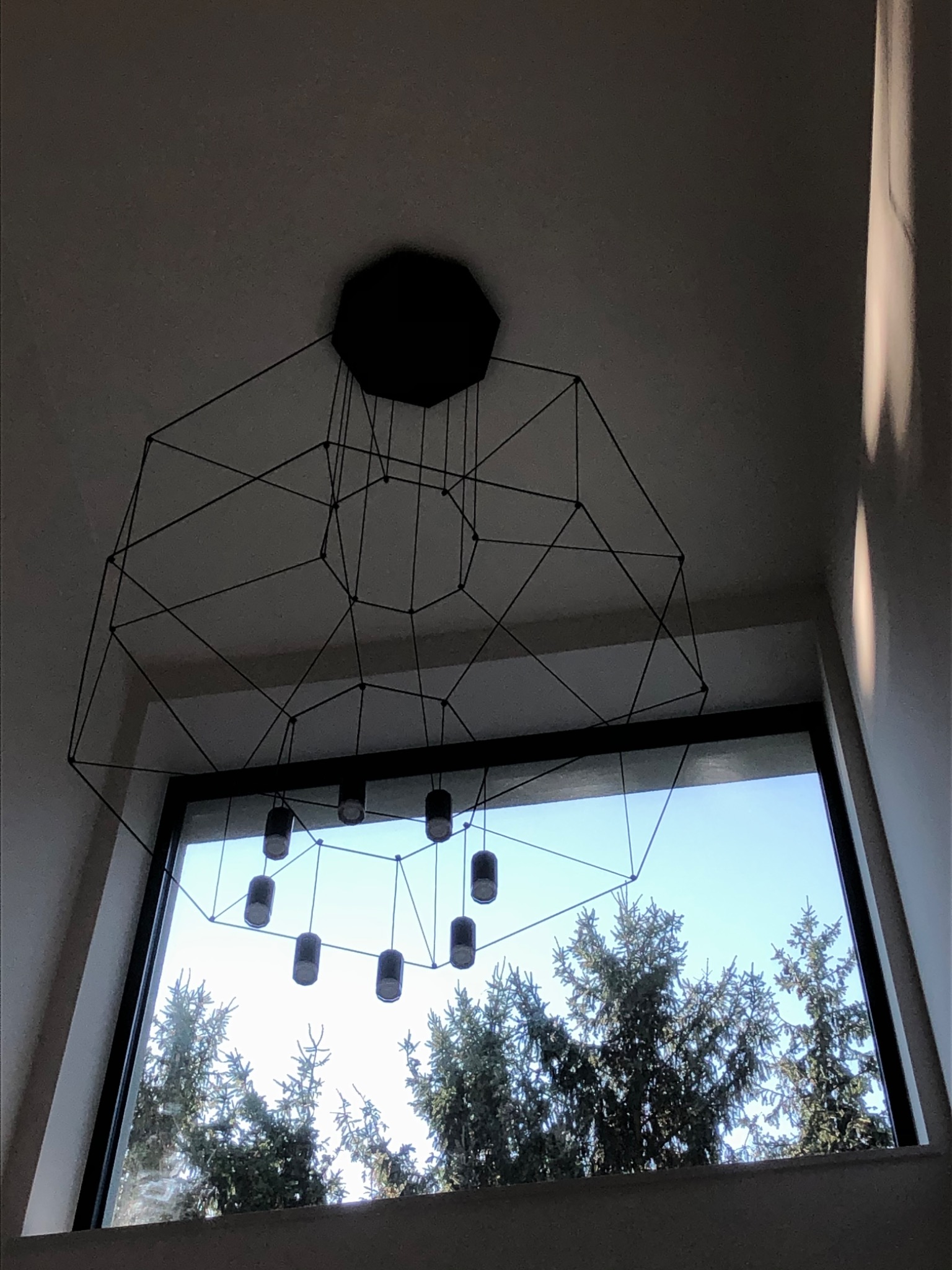 Geometryczna lampa wisząca z cylindrycznymi kloszami na tle okna z widokiem na drzewa iglaste i błękitne niebo.