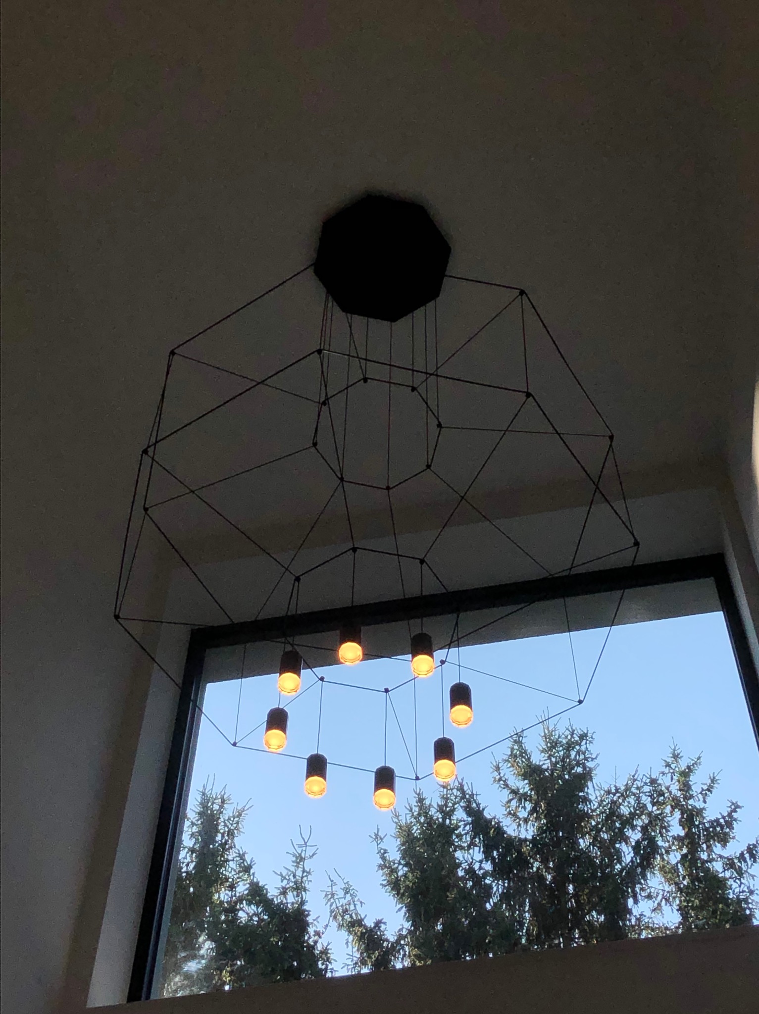 Nowoczesny żyrandol z czarnymi cylindrycznymi lampami i geometryczną konstrukcją na tle okna z widokiem na drzewa i błękitne niebo.