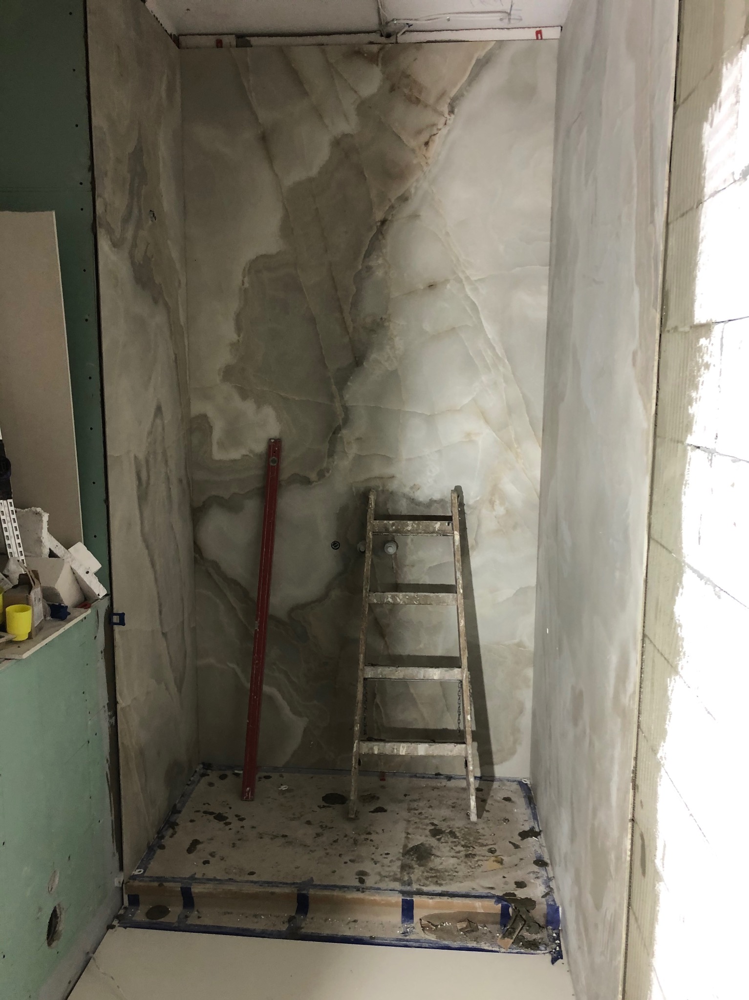 Fragment remontowanej łazienki z marmurowymi płytami na ścianach, poziomica oparta o ścianę, drabina w rogu i widoczne ślady zaprawy na podłodze.