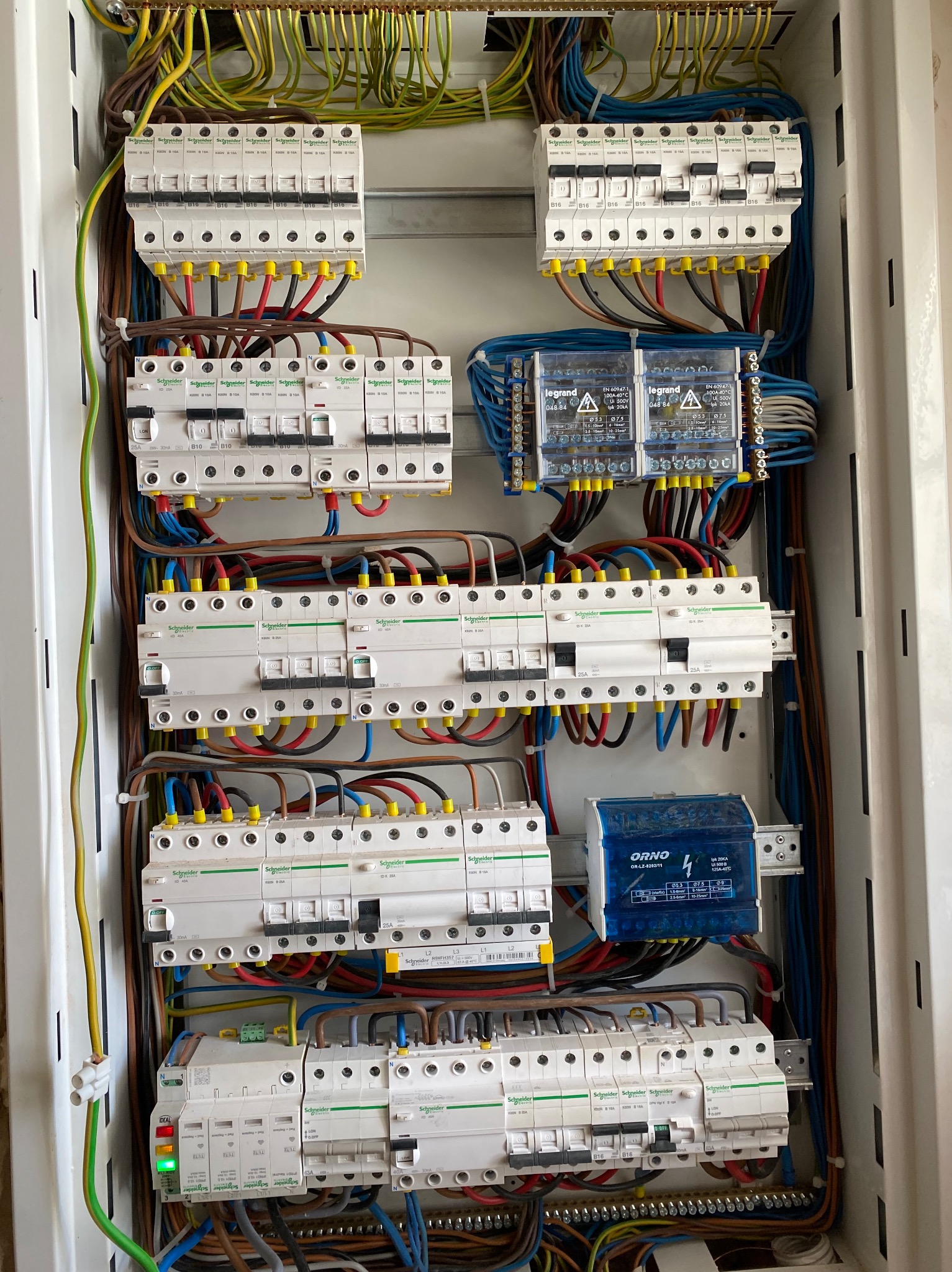 Otwarta skrzynka elektryczna z bezpiecznikami, przewodami i elementami sterującymi różnych marek, w tym Schneider Electric i Legrand, ułożonymi w uporządkowany sposób.