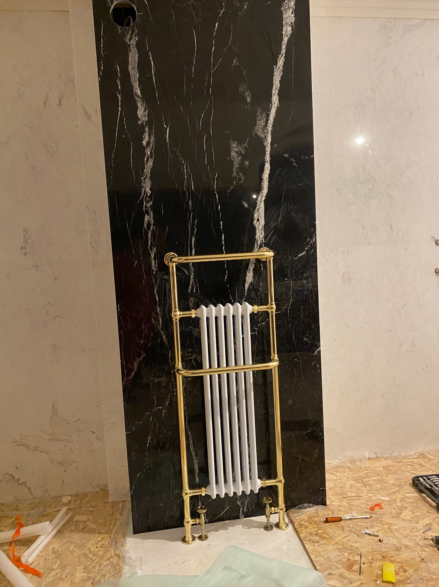 Montaż eleganckiego, złotego grzejnika łazienkowego na tle czarnej płyty z białym marmurkowym wzorem, podczas remontu łazienki.
