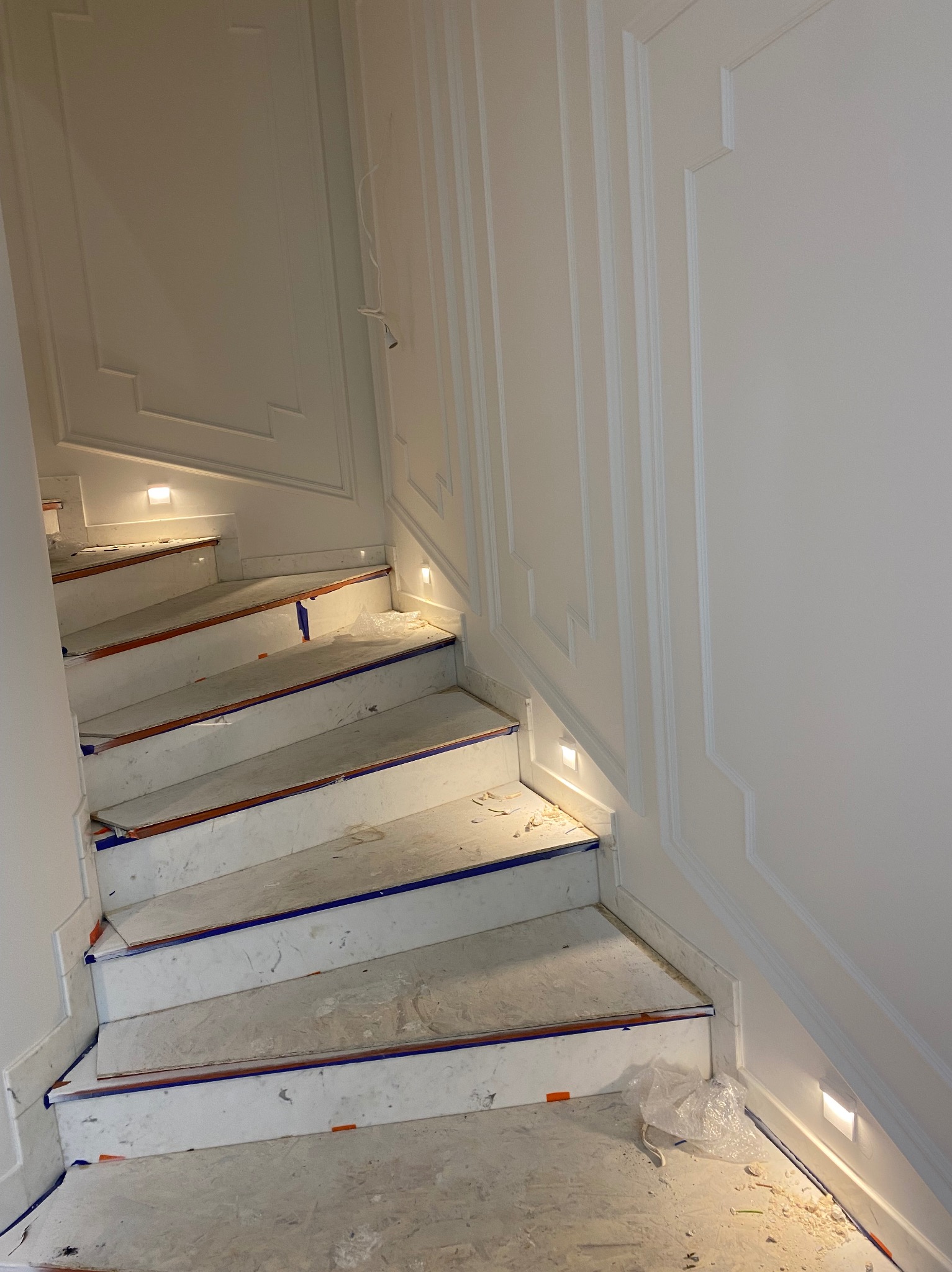 Klatka schodowa w trakcie prac wykończeniowych; widoczne białe ściany z dekoracyjnymi listwami, schody zabezpieczone taśmą malarską i folią, oraz wbudowane oświetlenie punktowe.