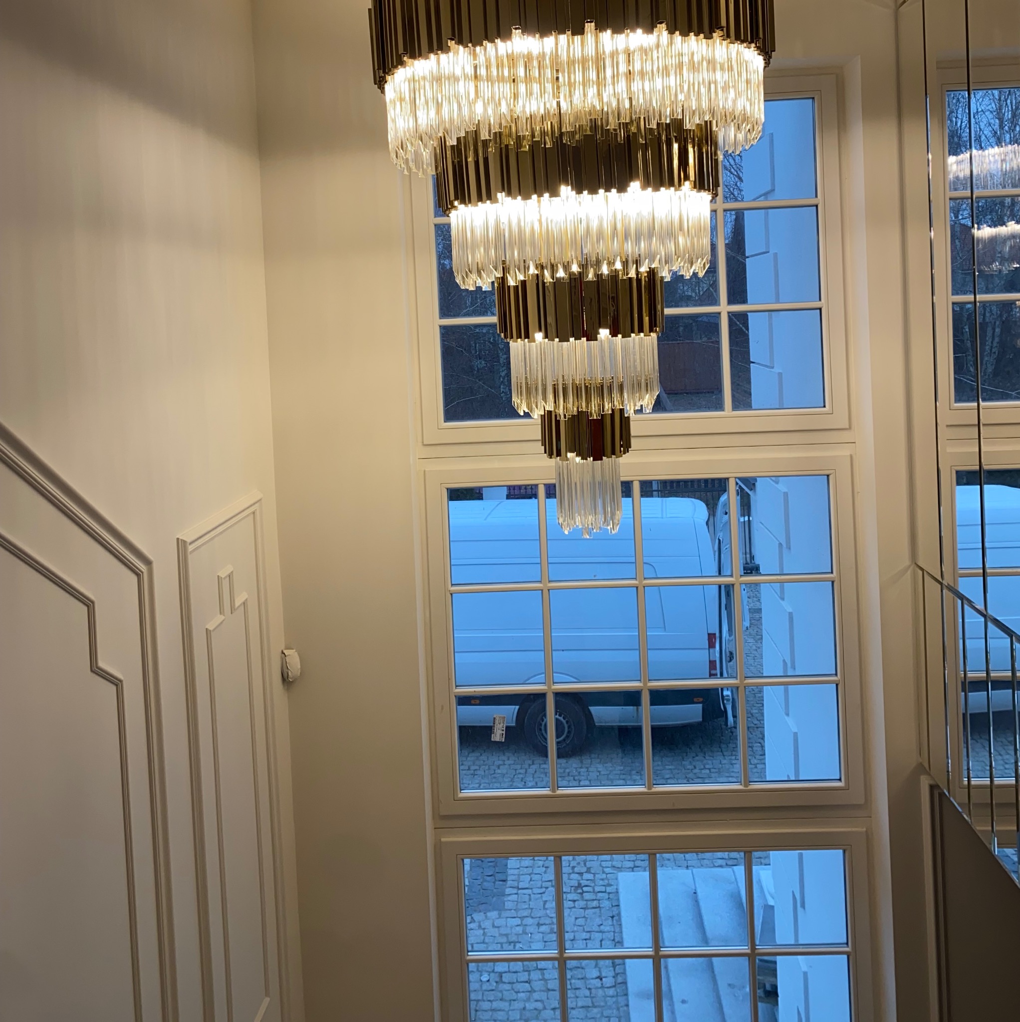 Wnętrze klatki schodowej z widokiem na wielopoziomowy, kryształowy żyrandol w stylu glamour, białe ściany z dekoracyjnymi listwami i widok z okna na biały samochód dostawczy zaparkowany...