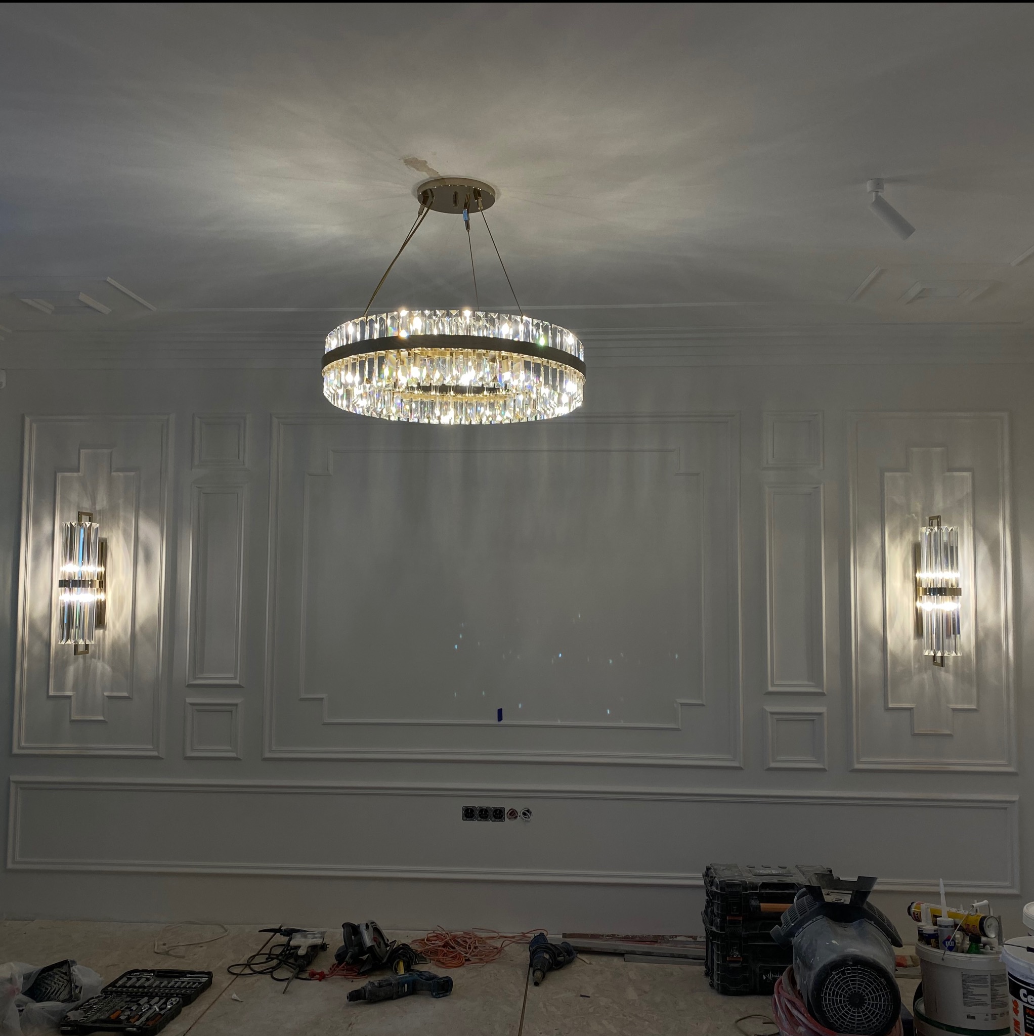Kryształowy okrągły żyrandol pod sufitem z dekoracyjnymi listwami, na ścianie kinkiety w stylu art deco, widoczne narzędzia i materiały budowlane na podłodze.