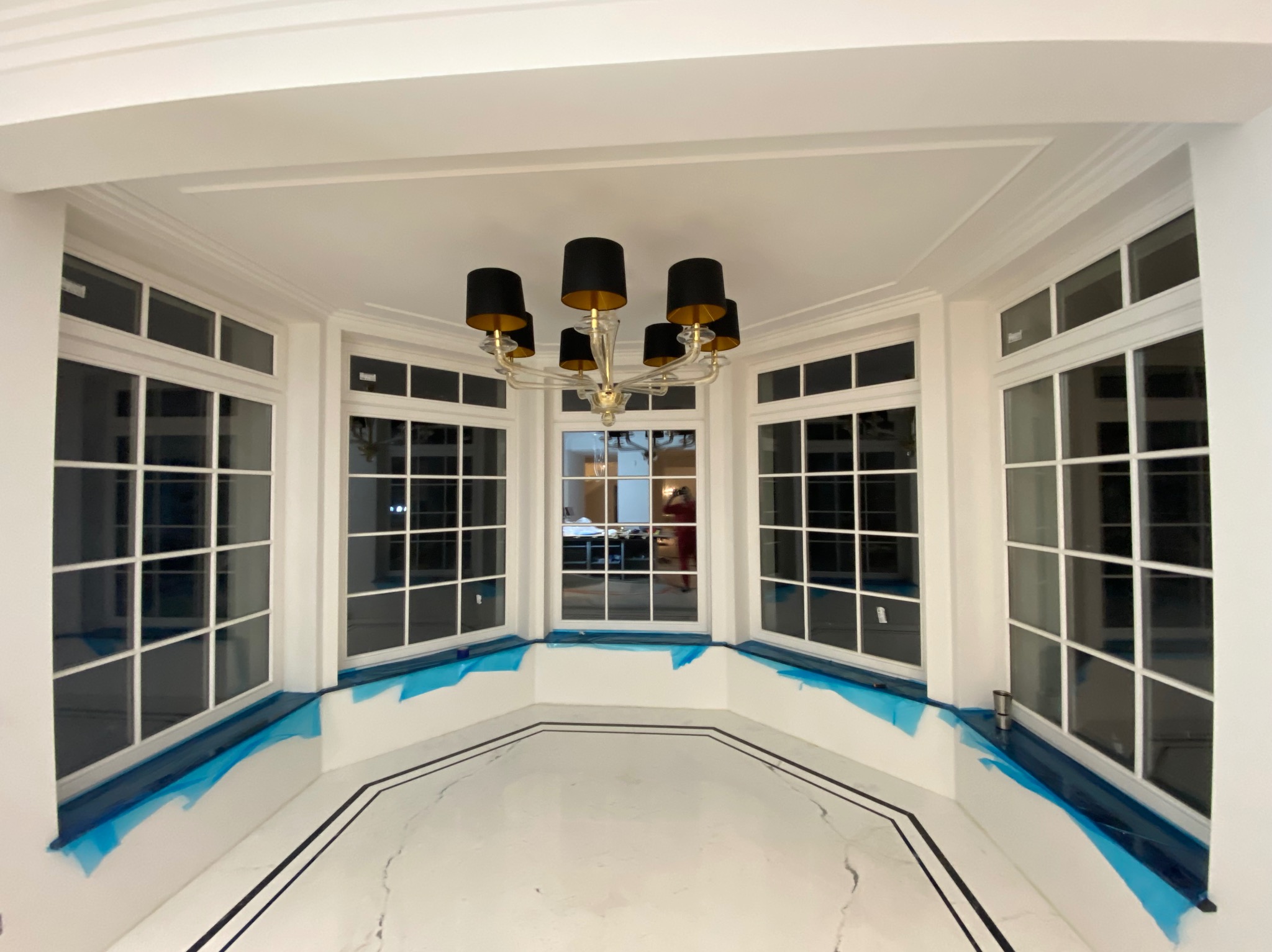 Jasne, przestronne wnętrze z wykuszem okiennym zabezpieczonym niebieską folią, marmurowa podłoga z czarnymi żyłami i nowoczesny żyrandol z czarnymi abażurami.