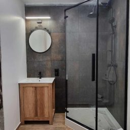 Kordian Kujawski - Nowoczesna łazienka w trakcie remontu: kabina prysznicowa z czarną ramą, drewniana szafka umywalkowa z okrągłym lustrem, płytki imitujące kamień na ścianach, drewnopodobne panele na podłodze...