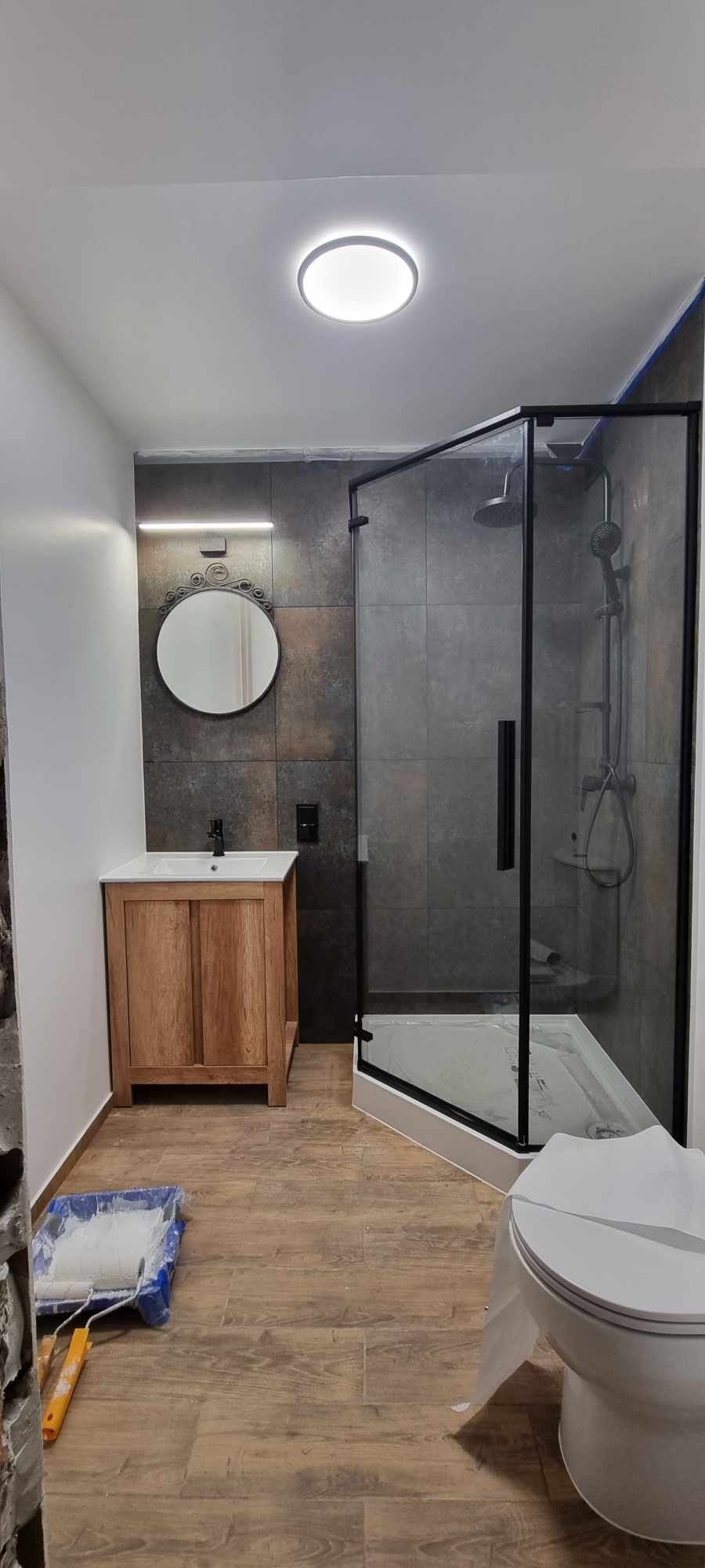 Nowoczesna łazienka w trakcie remontu: kabina prysznicowa z czarną ramą, drewniana szafka umywalkowa z okrągłym lustrem, płytki imitujące kamień na ścianach, drewnopodobne panele na podłodze...
