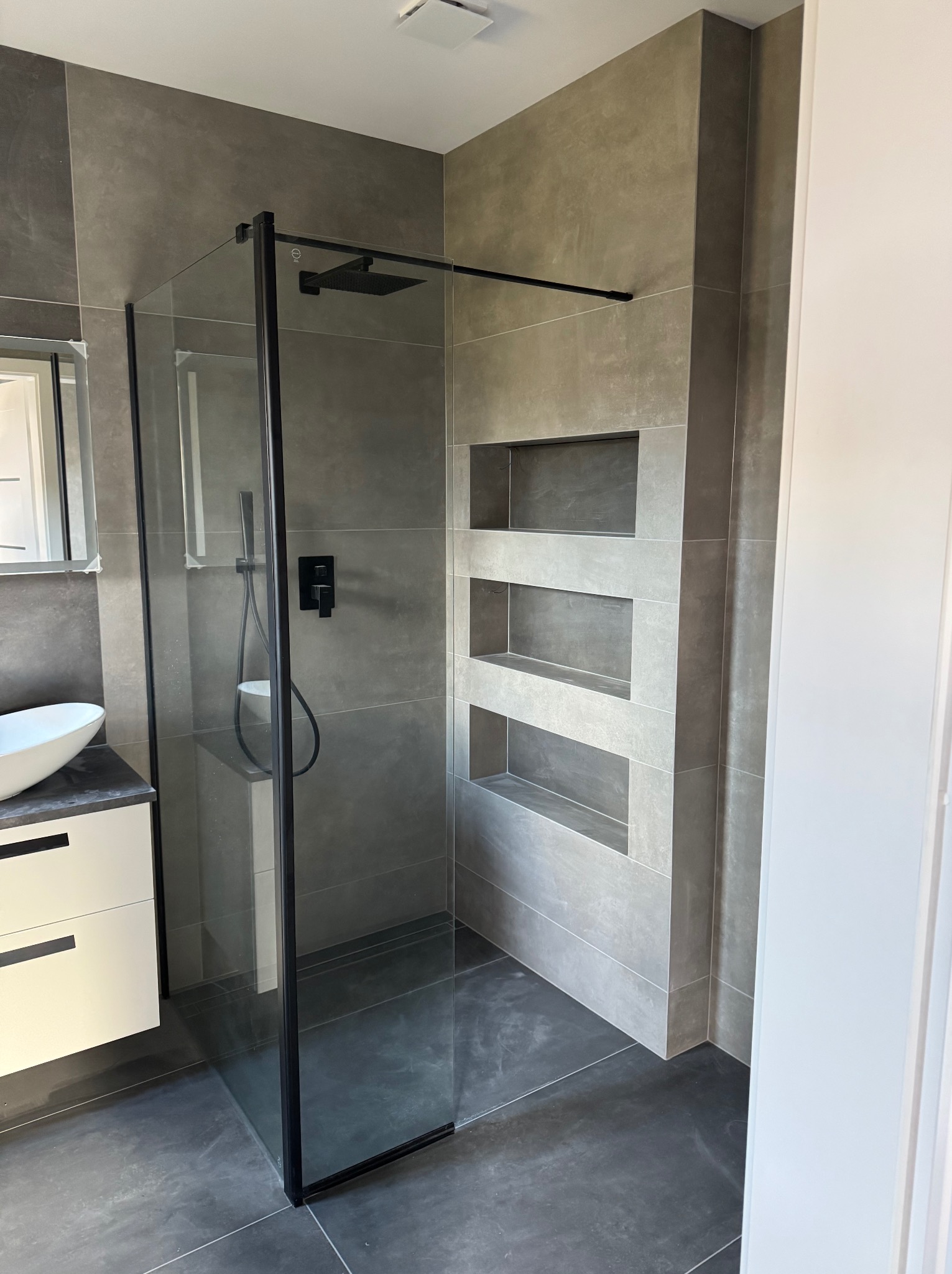 Nowoczesna łazienka z kabiną prysznicową typu walk-in z czarną ramą, szare płytki wielkoformatowe, wnęki w ścianie wyłożone płytkami, biała umywalka nablatowa na szafce z szufladami.