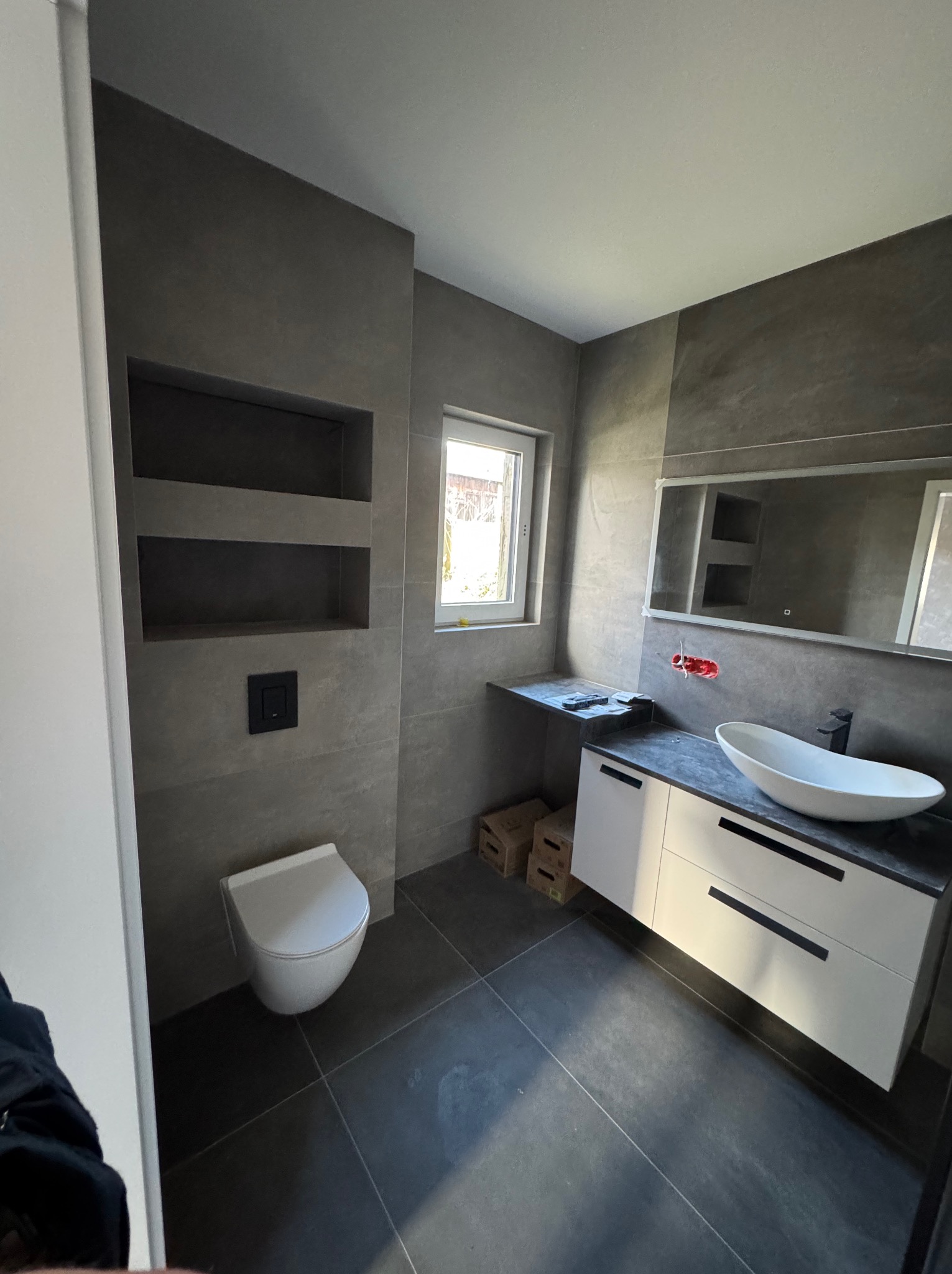 Nowoczesna łazienka z szarymi płytkami, podwieszaną toaletą, umywalką nablatową na białej szafce z czarnymi uchwytami, wnęką z półkami i dużym lustrem z podświetleniem.