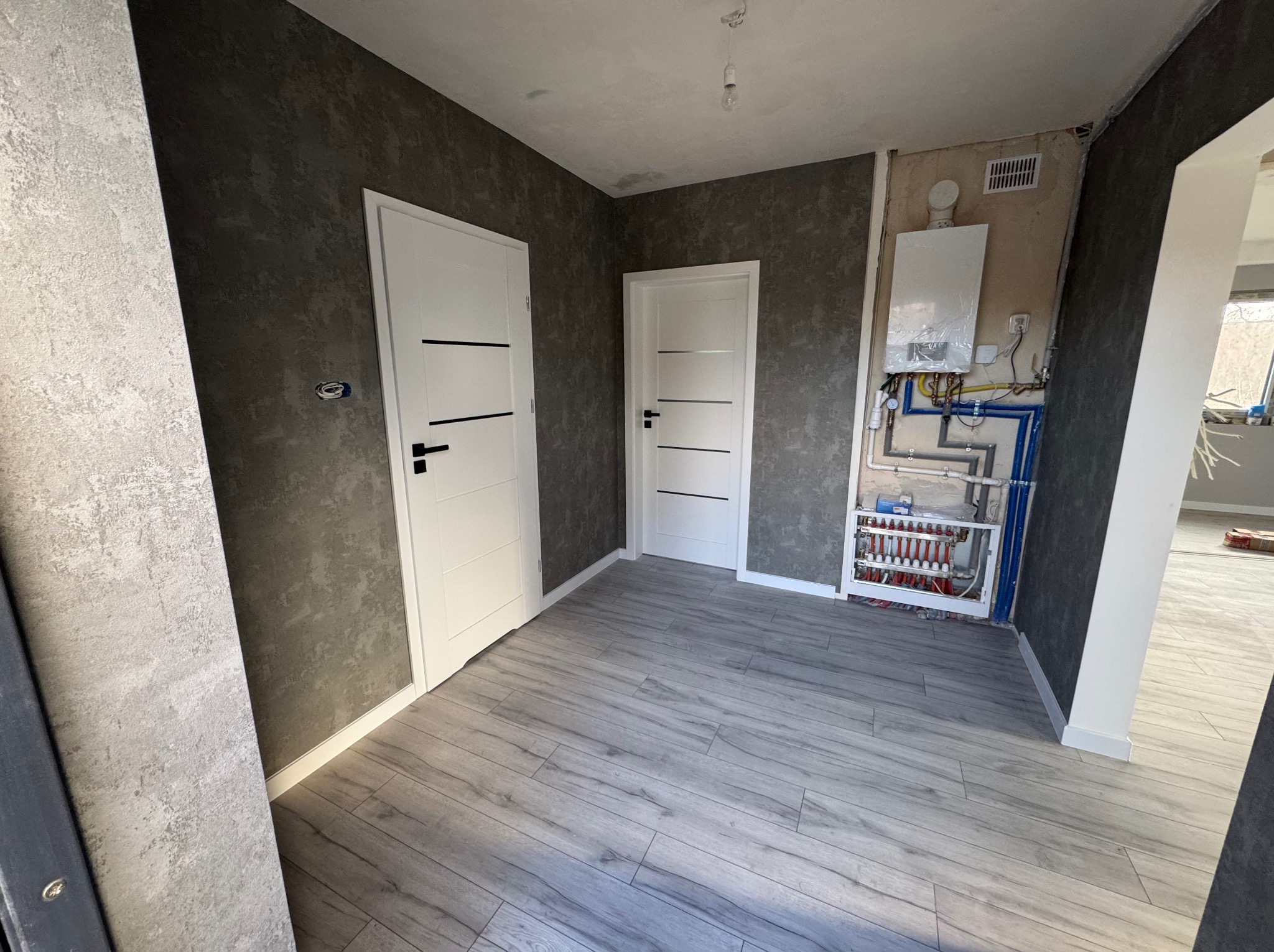 Wnętrze w trakcie prac wykończeniowych: dwie białe drzwi z czarnymi klamkami, szare panele podłogowe imitujące drewno, ściany o fakturze betonu w dwóch odcieniach szarości, widoczny fragment...