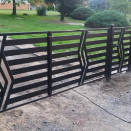 Nowoczesna, czarna, metalowa brama wjazdowa z poziomymi szczeblami i geometrycznym wzorem, widoczna na tle zielonego trawnika i drzew, z kablem leżącym na żwirowym podłożu.