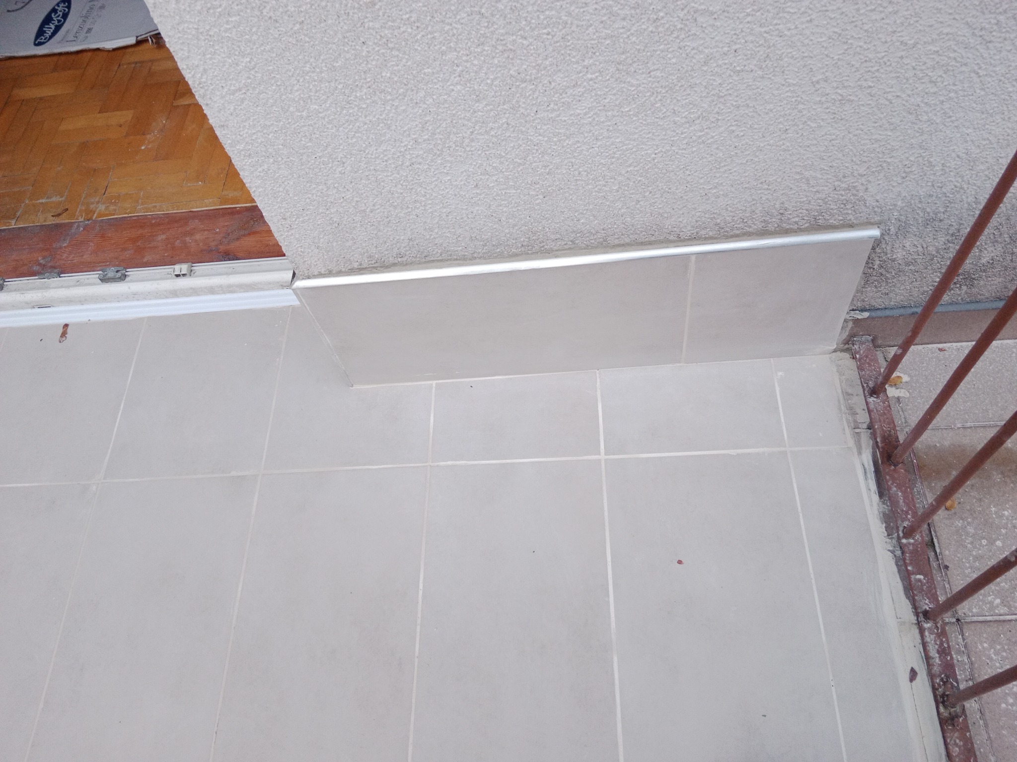 Jasne płytki podłogowe na balkonie, widoczna listwa przypodłogowa z metalowym wykończeniem, fragment starej drewnianej podłogi wewnątrz pomieszczenia, oraz fragment barierki balkonowej.