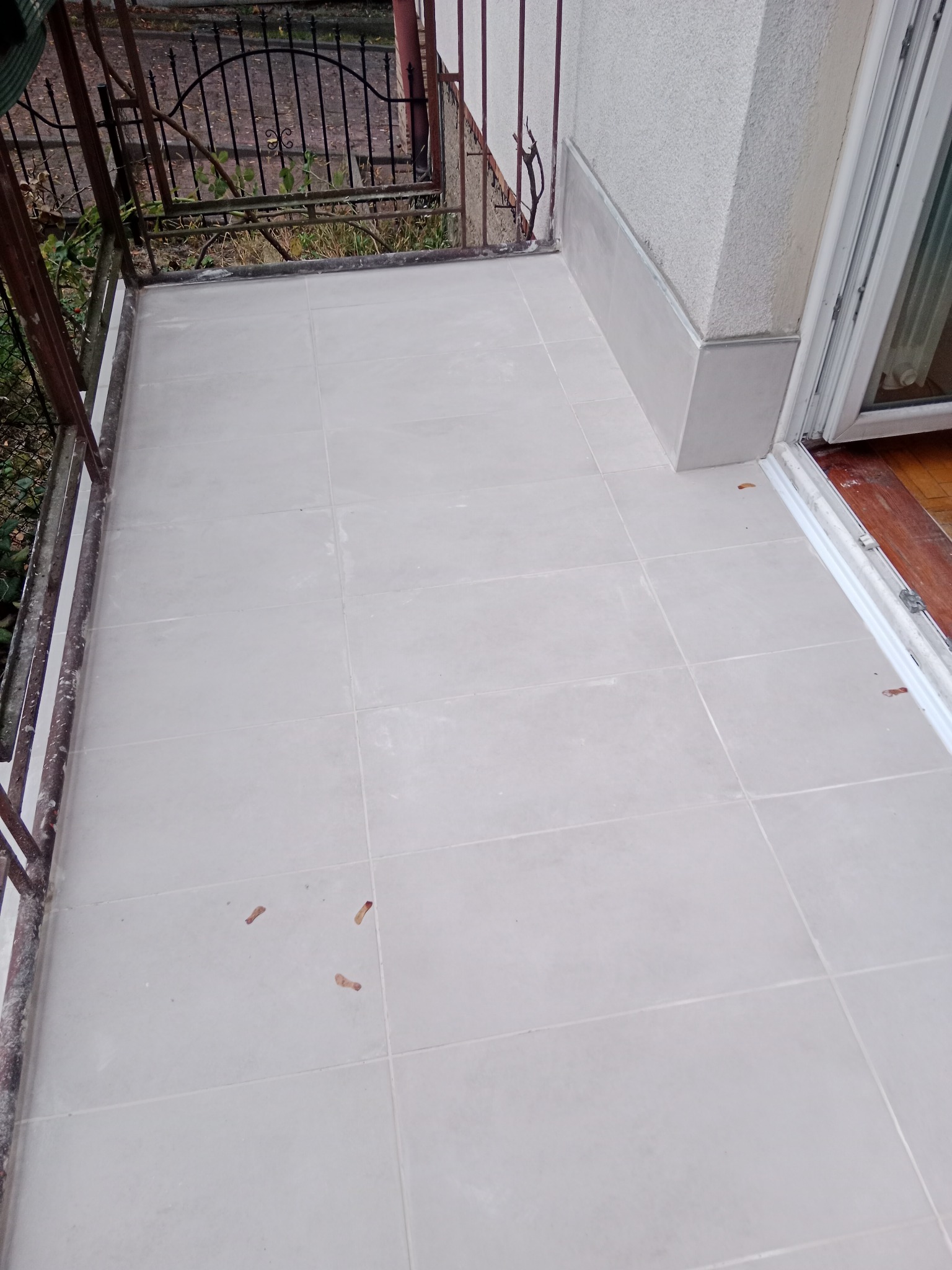 Jasnoszare płytki ceramiczne ułożone na balkonie, widoczne ślady zaprawy, metalowa balustrada i fragment ściany z cokołem z płytek.