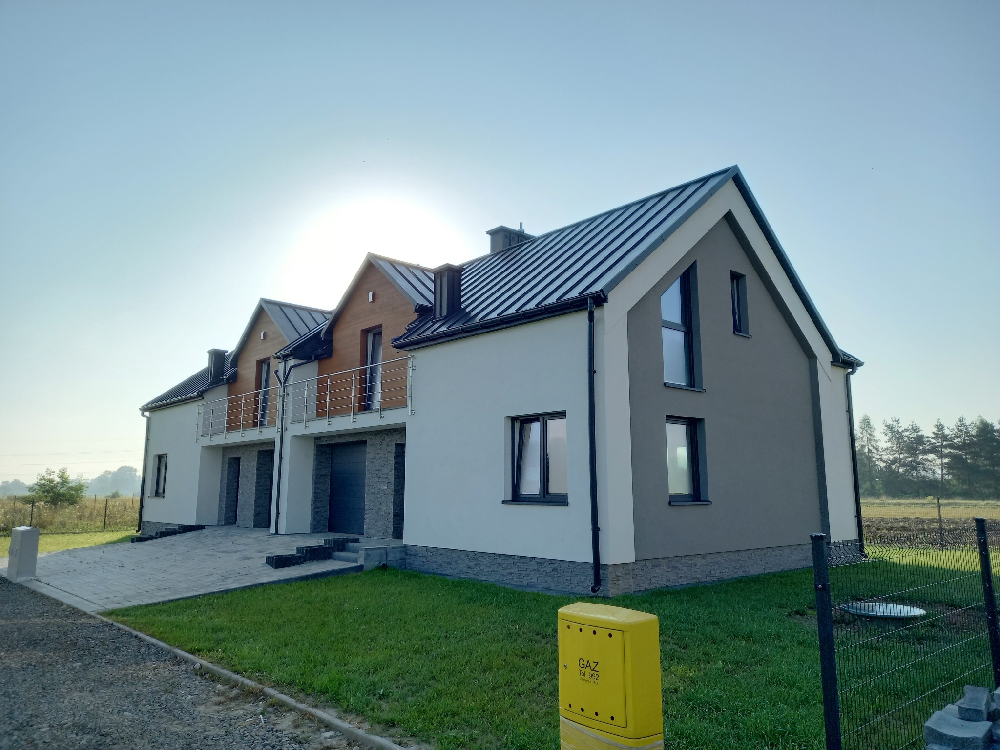 Nowoczesny bliźniak z elewacją w odcieniach bieli i szarości, z elementami drewnianymi, metalowy dach, kostka brukowa na podjeździe, skrzynka gazowa w kolorze żółtym.