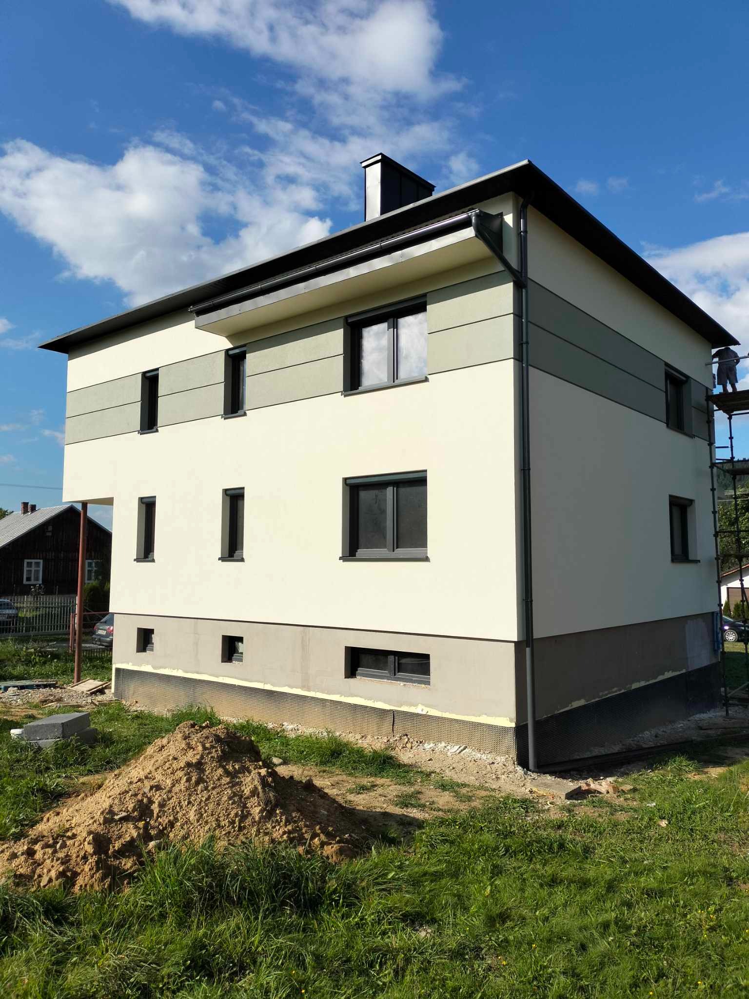 Dwukondygnacyjny dom jednorodzinny z elewacją w odcieniach beżu i szarości, z pracownikiem na rusztowaniu przy prawej ścianie, widoczne okna z czarnymi ramami, na pierwszym planie hałda ziemi.