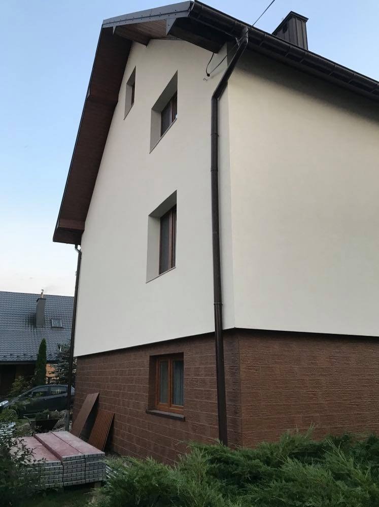 Wykończona elewacja domu jednorodzinnego z brązowym panelem imitującym drewno na dolnej części i jasną, gładką powierzchnią powyżej, widoczne okna i rynna.