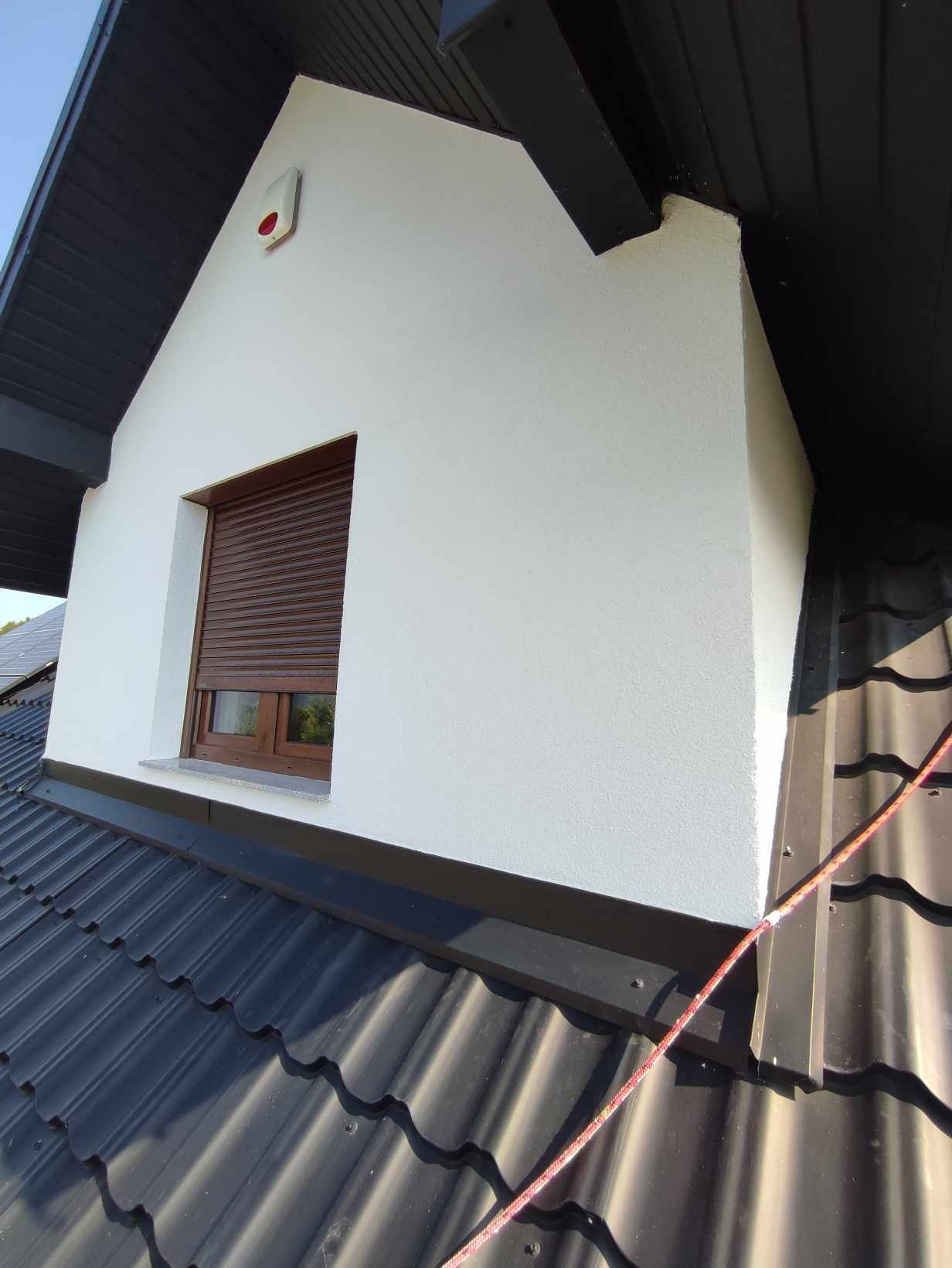 Poddasze domu z oknem i brązowymi roletami, ściana pokryta białą, chropowatą strukturą, widok z dachu pokrytego czarną blachodachówką, z boku widoczny czujnik alarmowy.