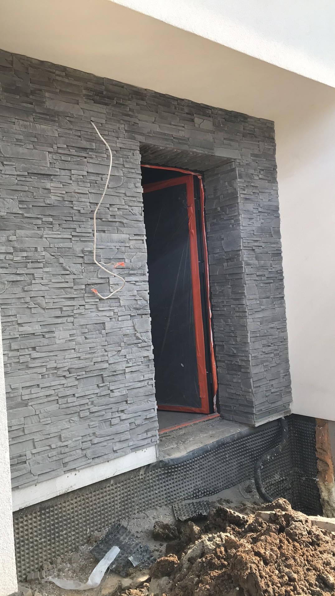 Elewacja wykończona szarą okładziną kamienną wokół otworu drzwiowego, zabezpieczonego folią; widoczna izolacja fundamentów i fragment przewodu elektrycznego.