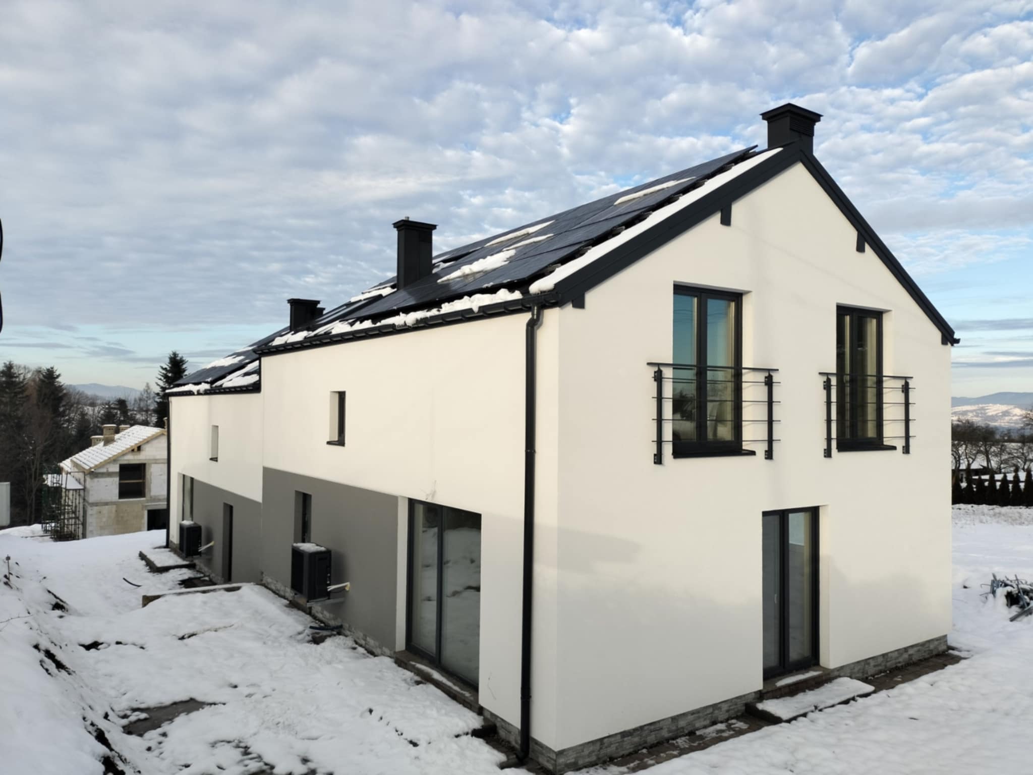 Nowoczesny, dwukondygnacyjny dom z białymi ścianami i czarnym dachem pokrytym panelami słonecznymi, widok z zewnątrz w zimowej scenerii, częściowo ośnieżony teren i drugi budynek w tle.