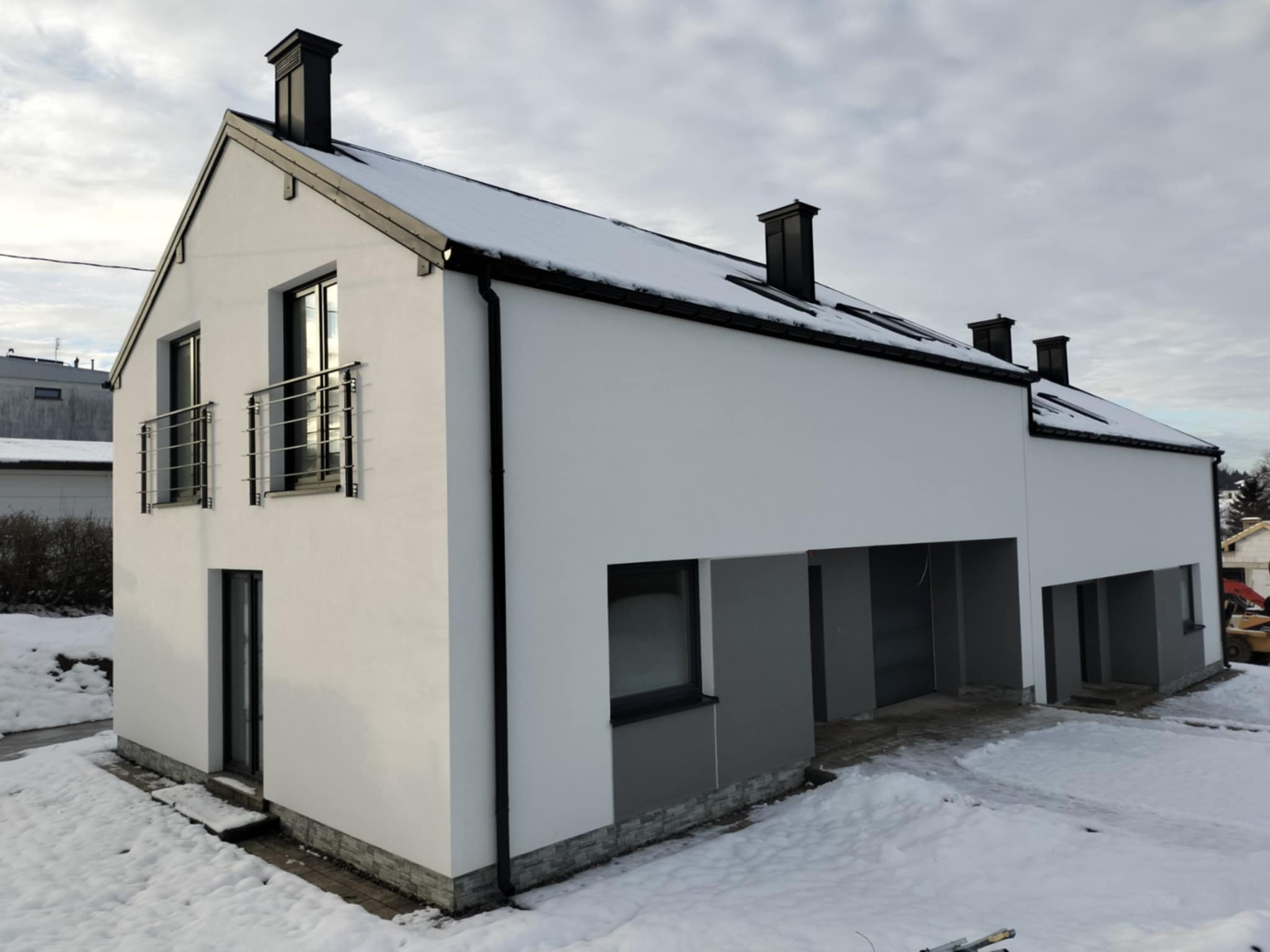 Nowoczesny, biały dom jednorodzinny z płaskim dachem i czarnymi kominami, częściowo pokryty śniegiem, z szarymi elementami wykończeniowymi elewacji i balkonem z metalową balustradą.